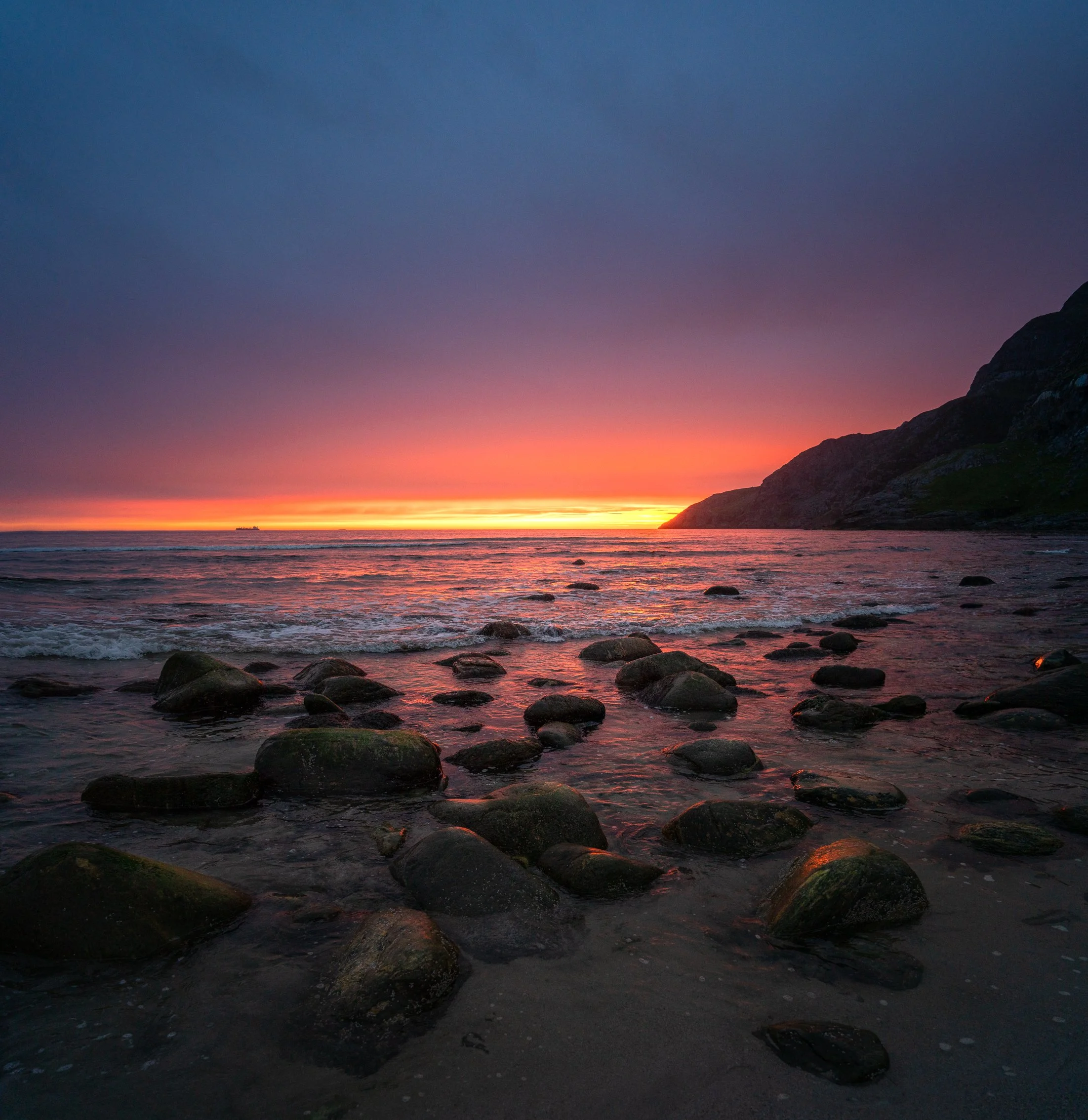A sunset over a rocky beach with dark clouds in the sky and ocean waves gently hitting the shore.