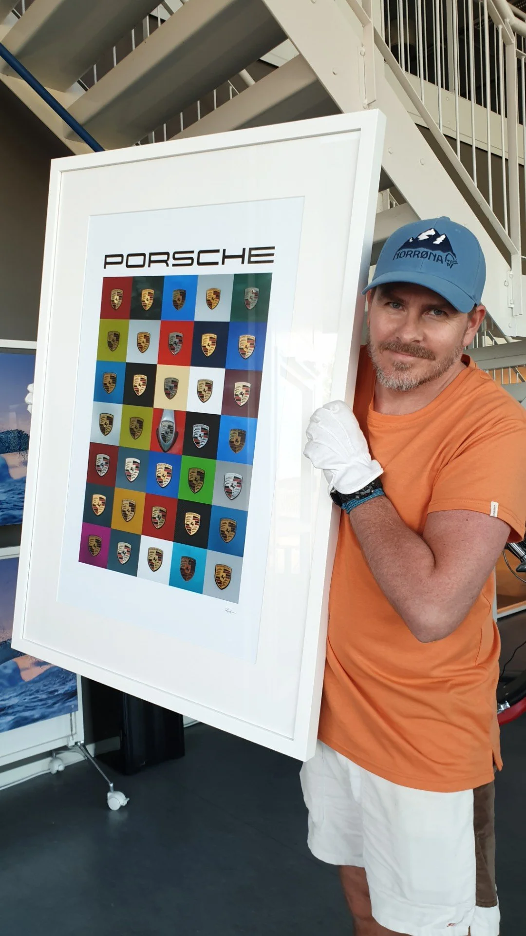 A man in an orange shirt and white gloves holding a framed print of various Porsche logos, standing inside a building with stairs in the background.