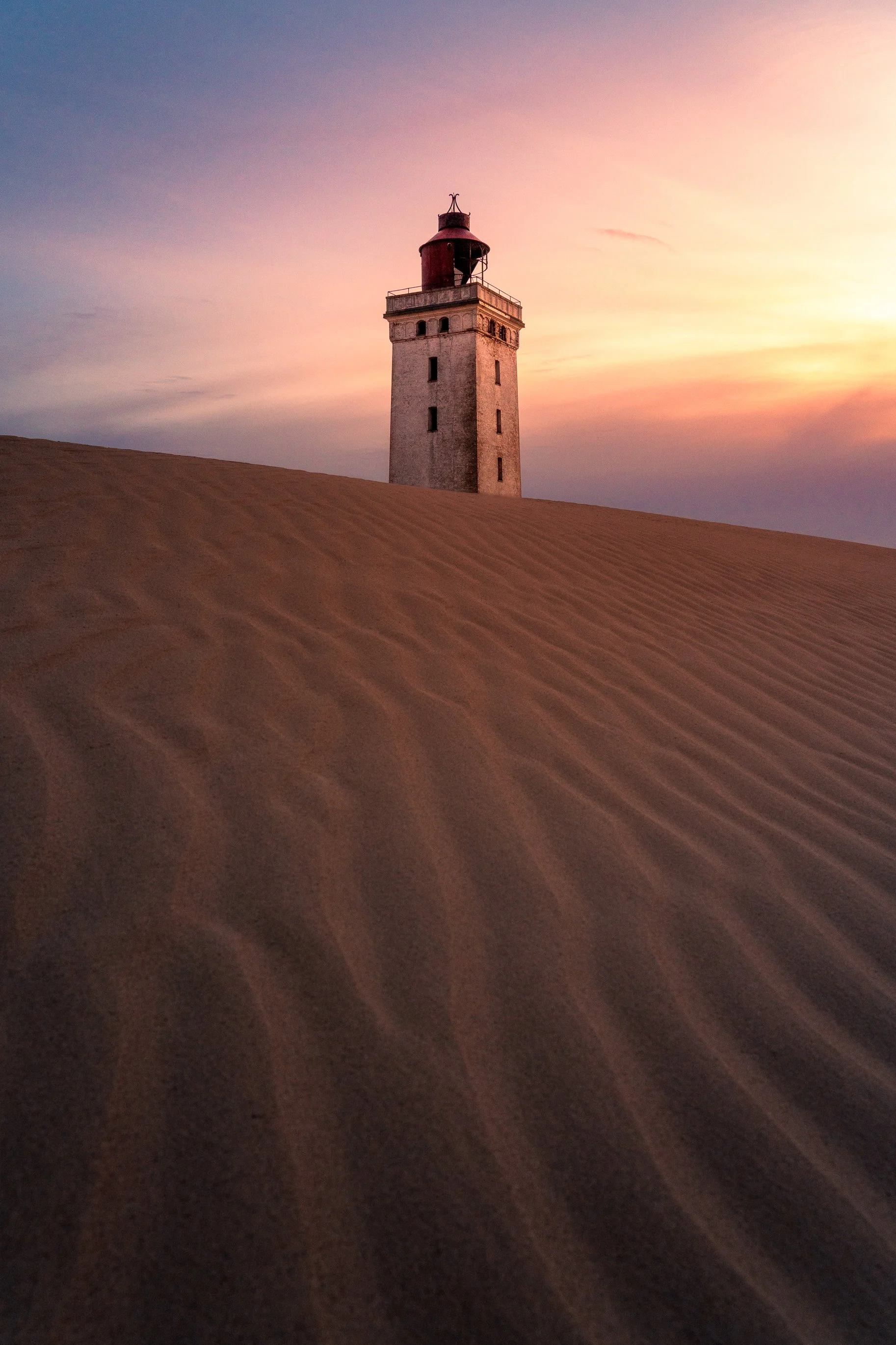 A lighthouse standing on a sand dune during sunset with a pink and purple sky.