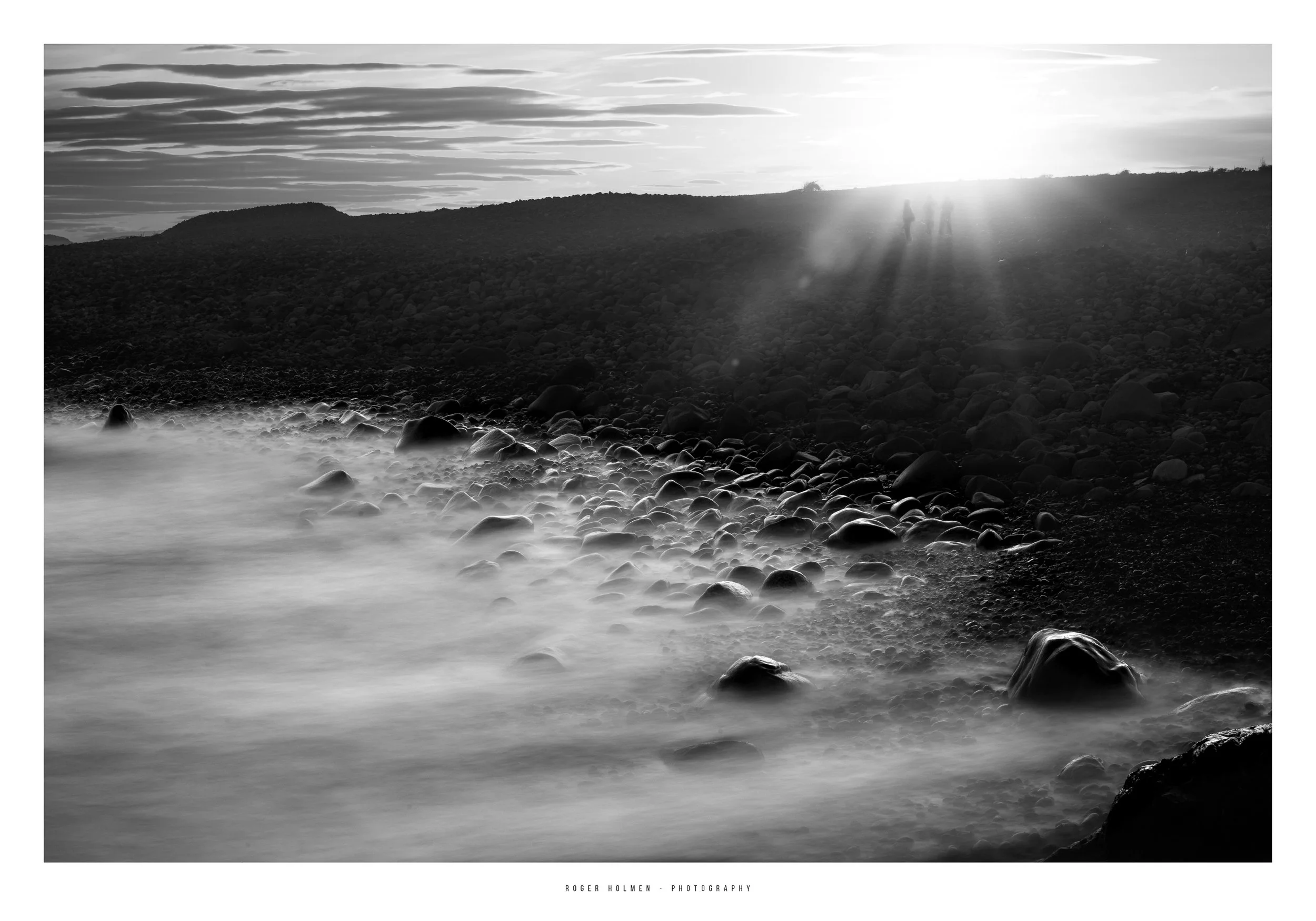 Black and white photo of a rocky shoreline with water flowing over stones, a hill in the background, and three people standing on the hill silhouetted by the setting sun.