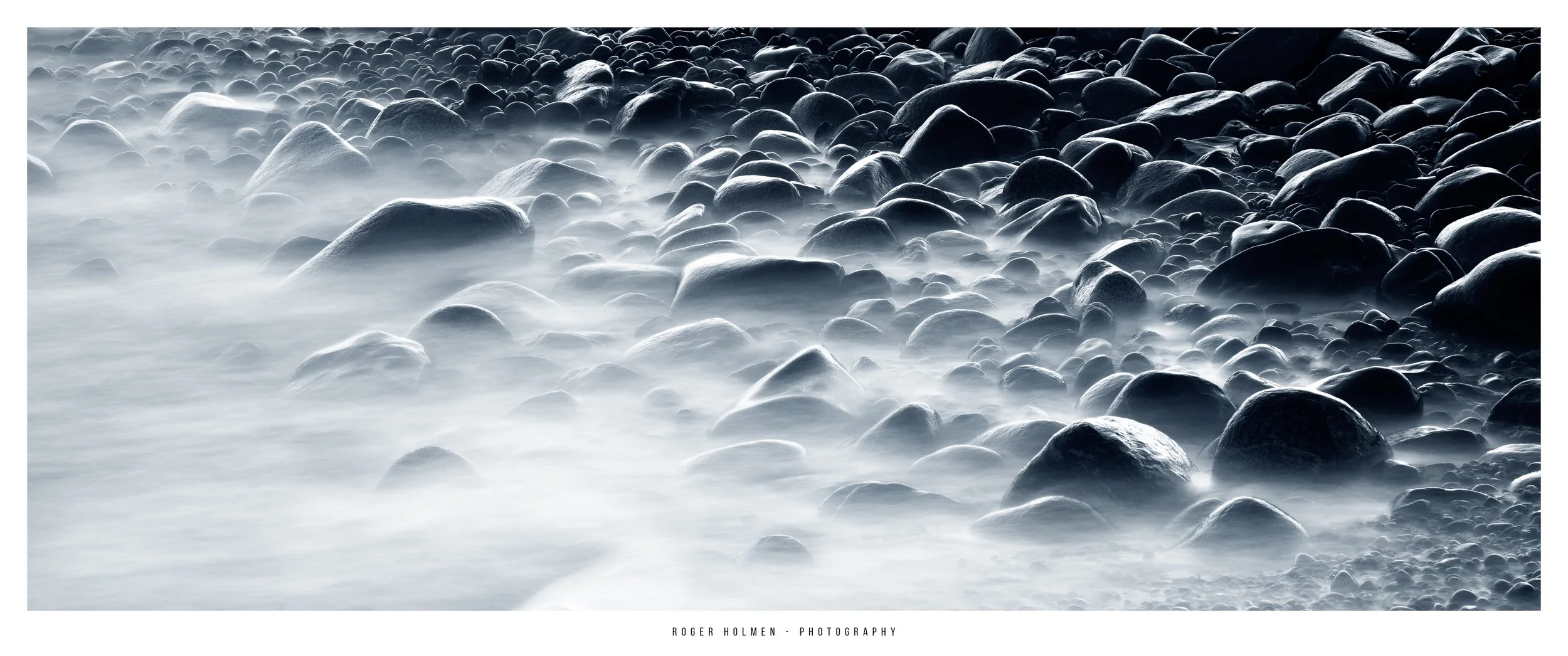 Black and white photograph of smooth stones and pebbles along a shoreline, with mist or fog intensifying the moody atmosphere.