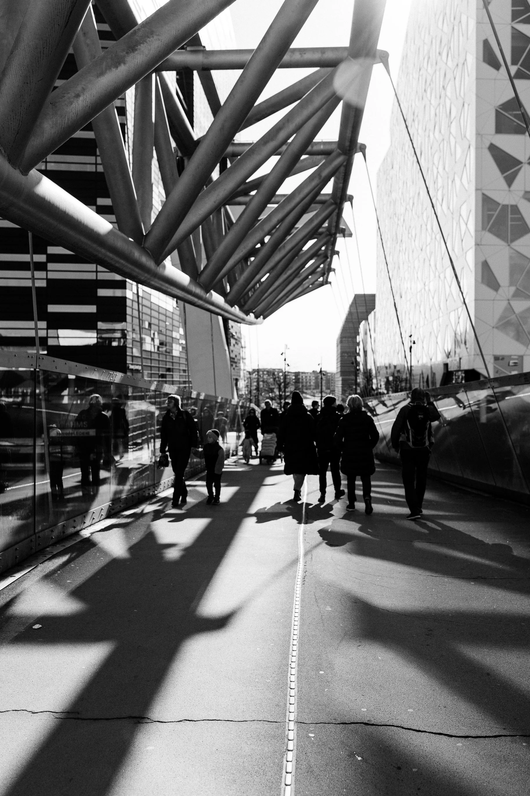 People walking along a modern urban street with shadows cast by architectural structures, black-and-white photo.