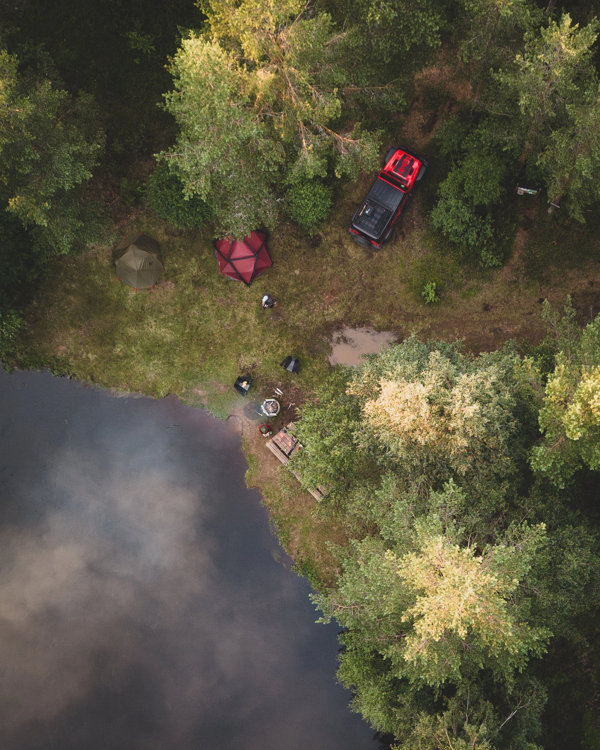 Aerial view of a lakeside camping site with two tents, a parked red and black truck, and a person near the lakeshore surrounded by trees.