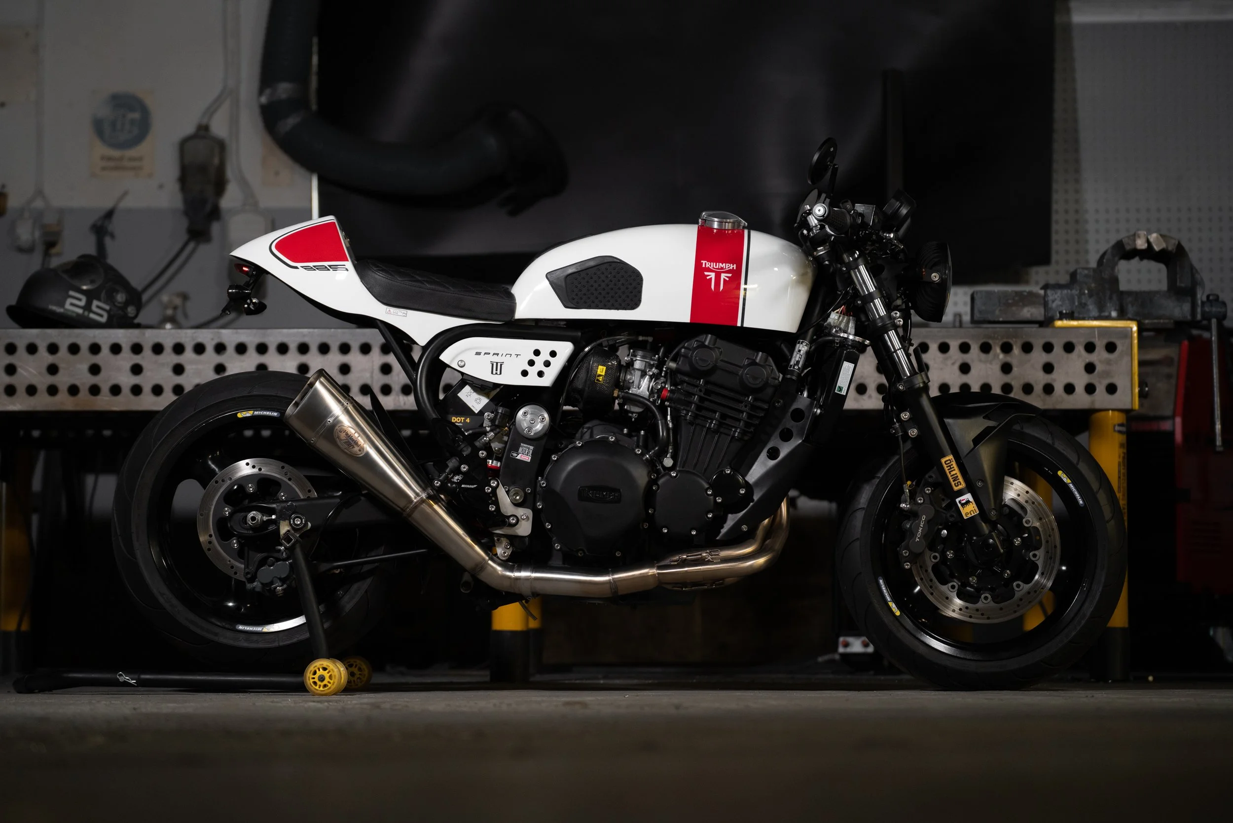 A Triumph sport motorcycle with a white and red fuel tank, black frame, and black tires, parked in a garage on a stand.