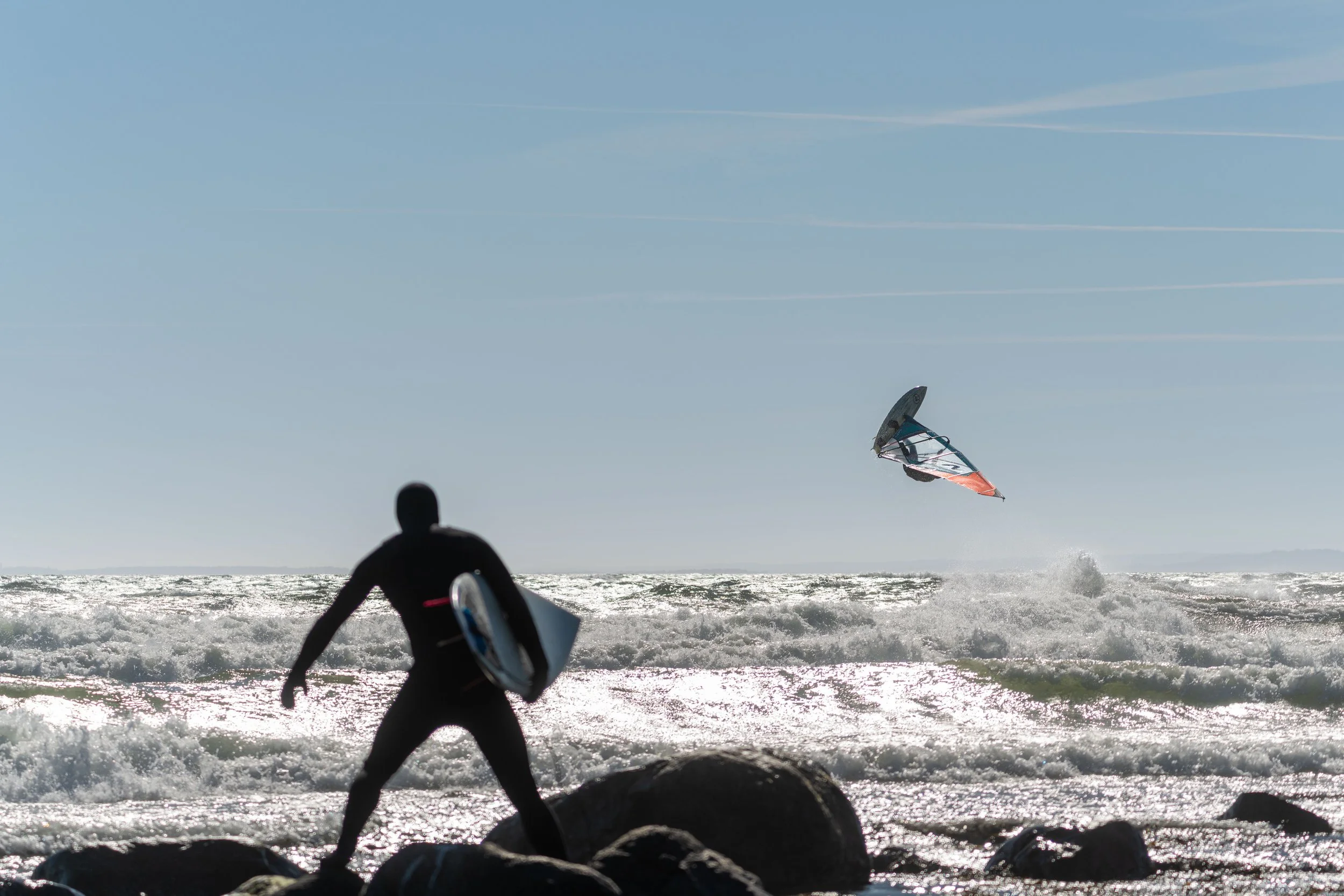 A person standing on rocks watching a windsurfer on the ocean. The windsurfer is airborne above the water with a sail. The sky is clear with some contrails. Location, Saltstein, Norway.