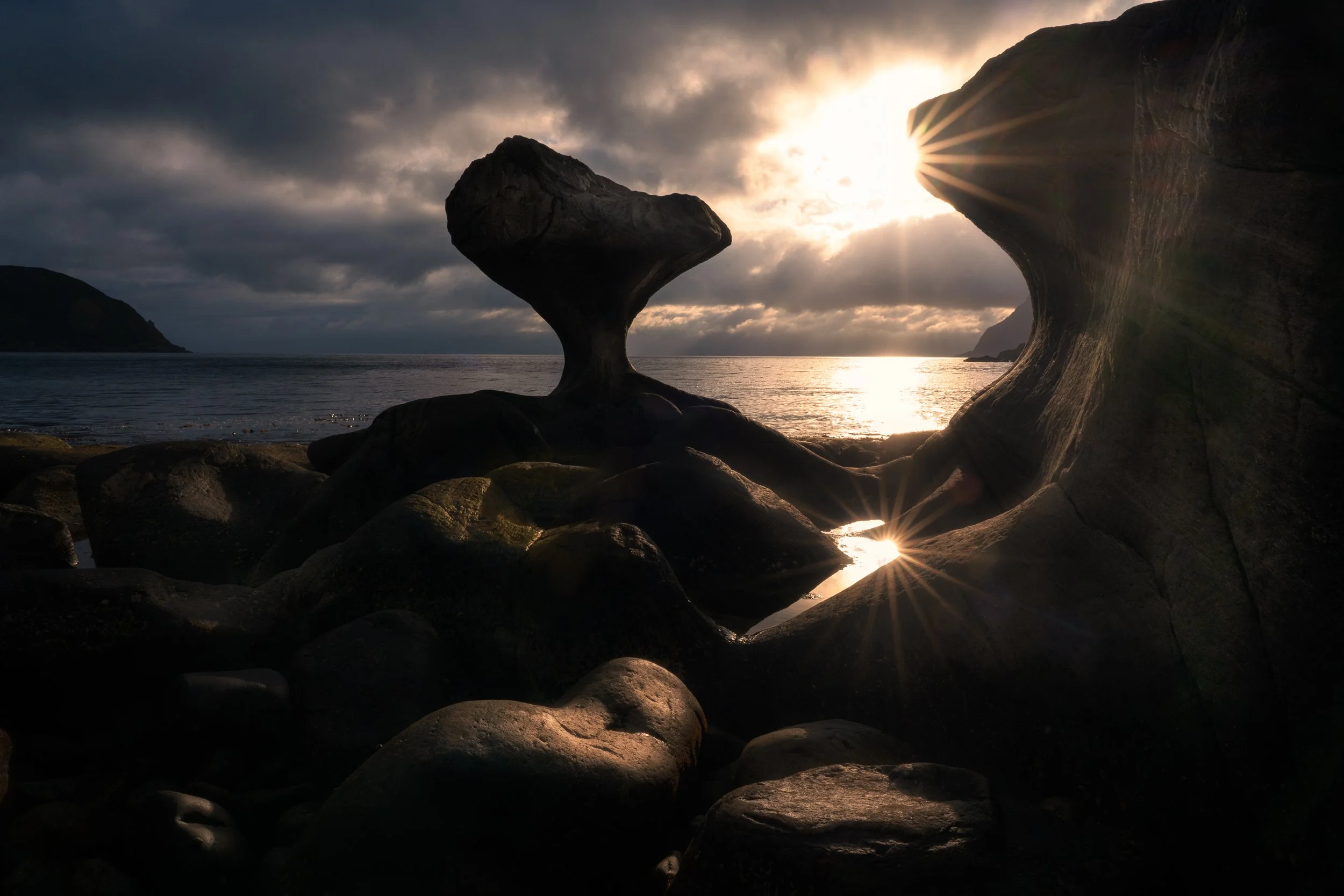 A rocky coastline at sunset with the sun’s rays creating starburst effects through a natural rock arch and a balanced rock formation.