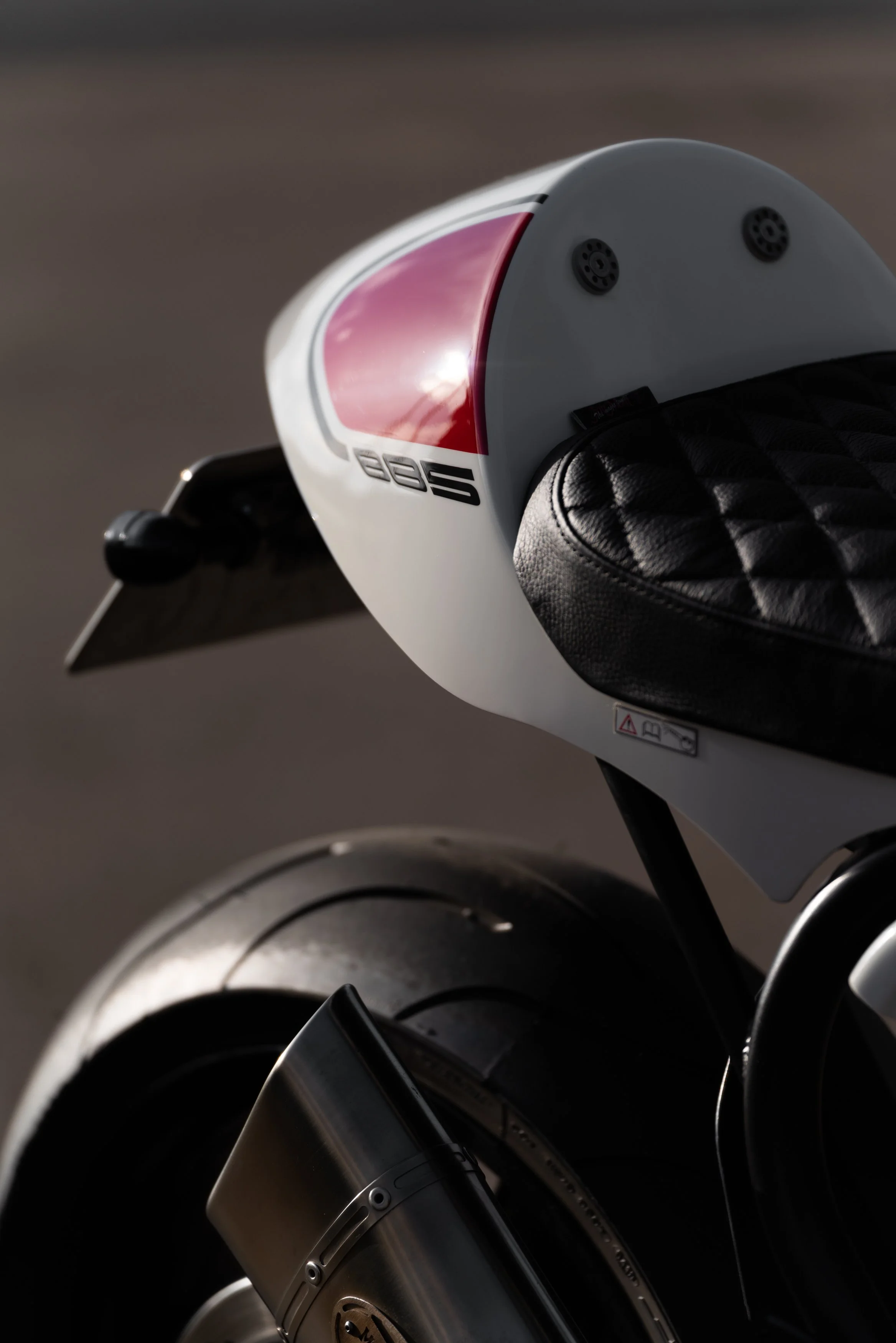 Close-up of a modern electric motorcycle with a white body and a black leather seat, focusing on the rear section including the tail light and exhaust details.