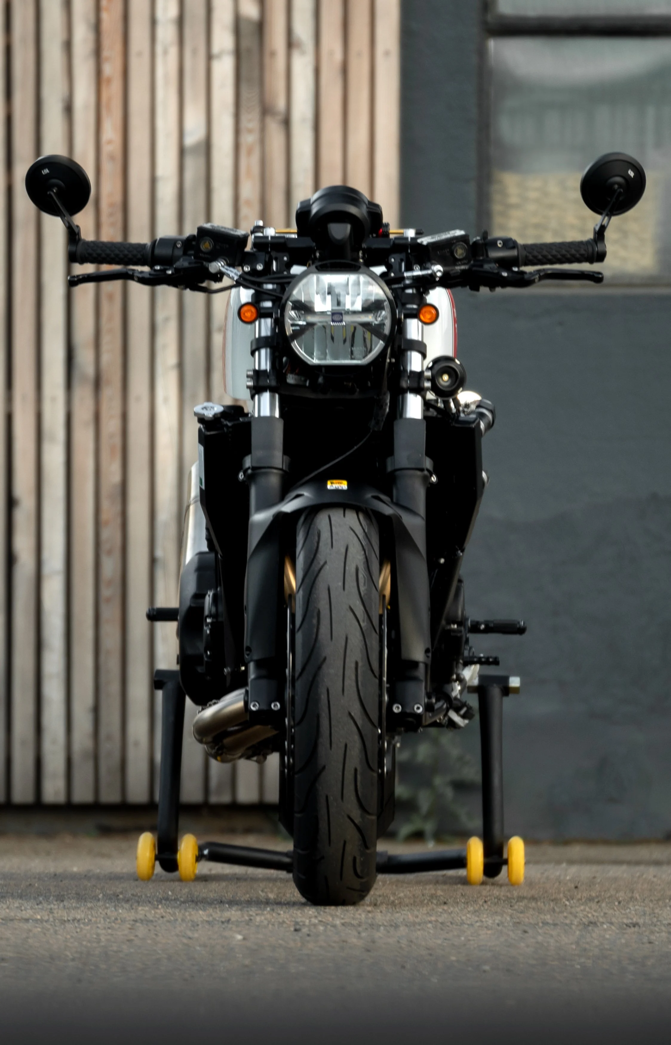 Front view of a black motorcycle with a single headlight, sitting on a stand with yellow wheels, against a background of a wooden fence and black wall.
