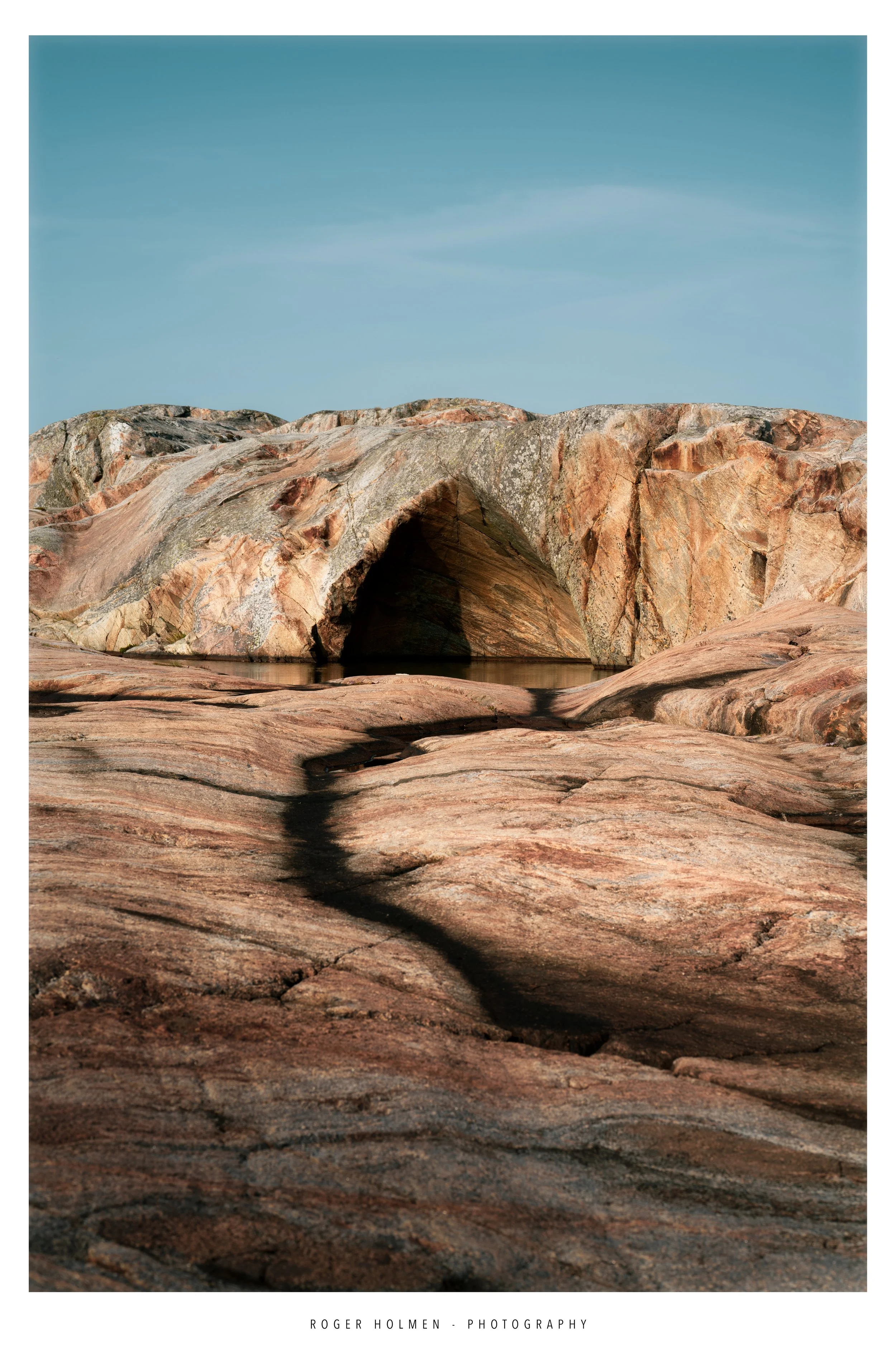 Rock formation with a natural arch, reflecting in a shallow water pool, under a clear blue sky.