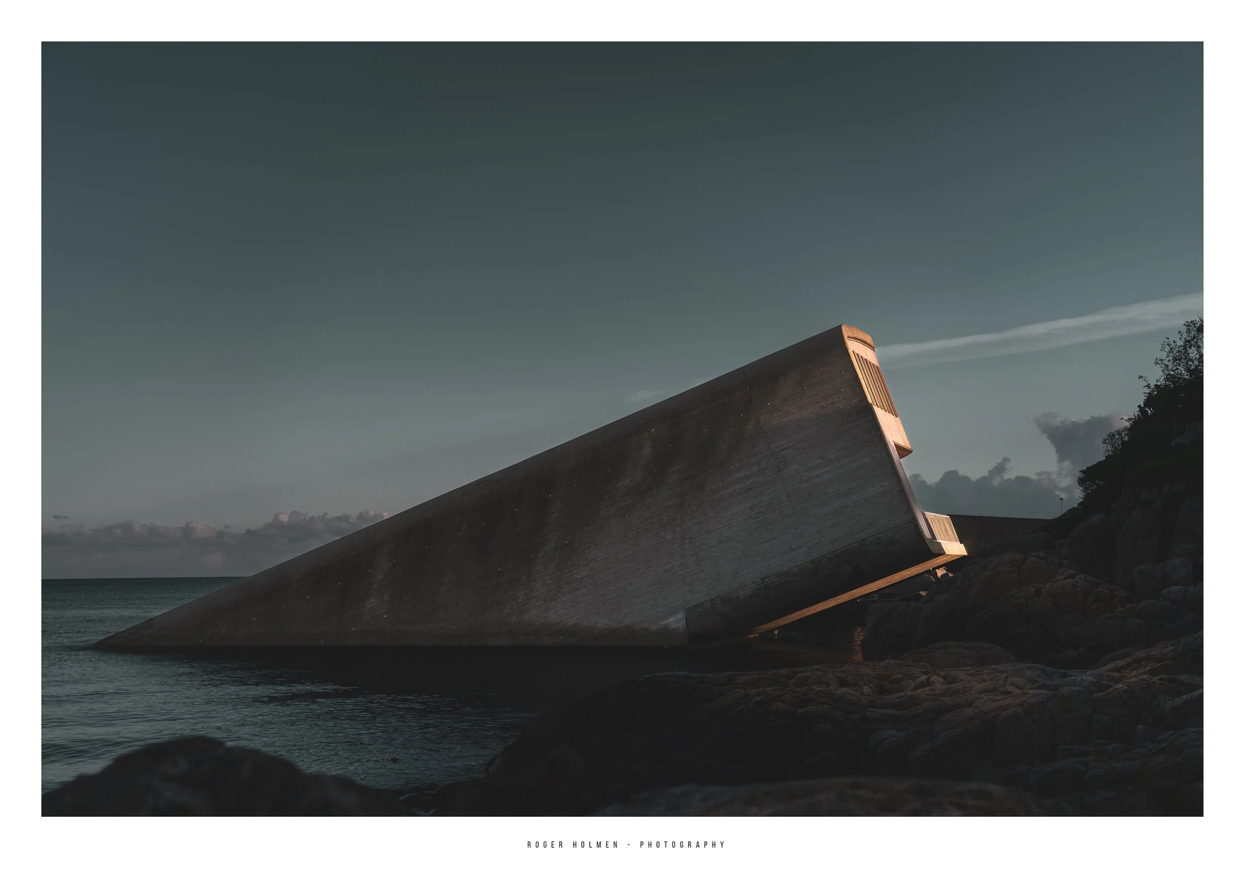 A large, tilted concrete structure on a rocky shoreline during twilight, with a cloudy sky and calm water in the background.