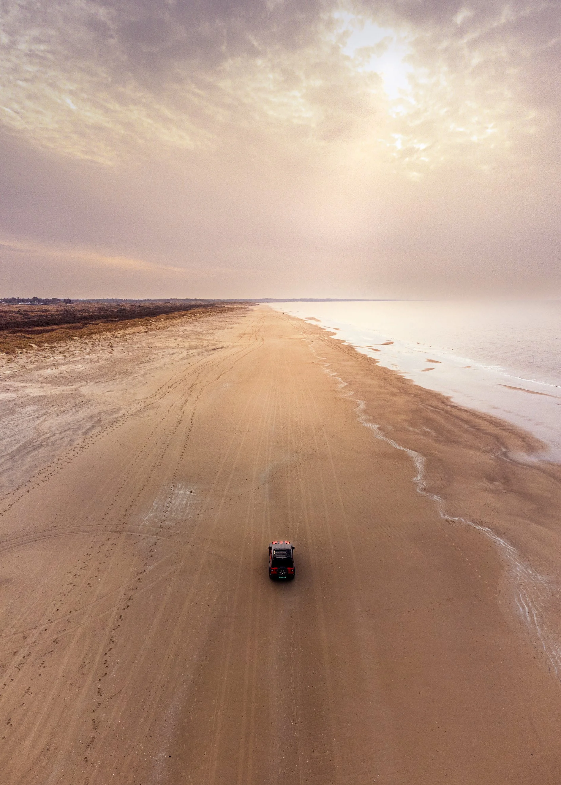 A lone vehicle driving on a sandy beach during daylight with cloudy skies.