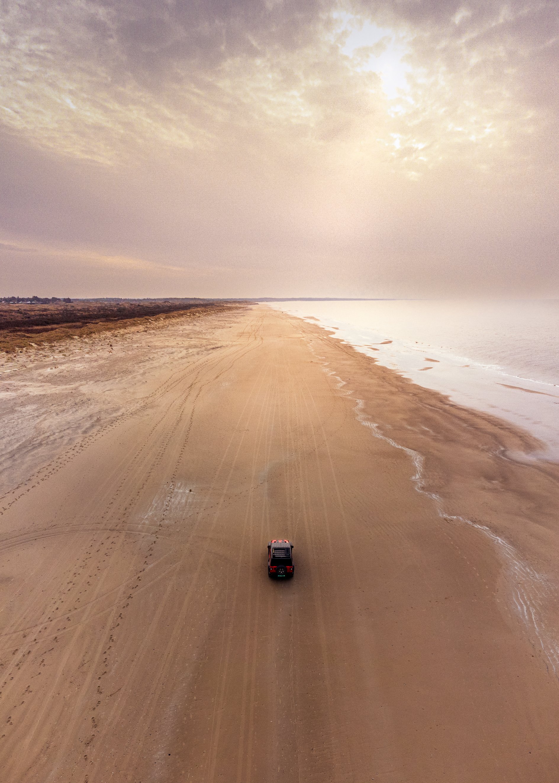 A lone vehicle driving on a sandy beach during daylight with cloudy skies.