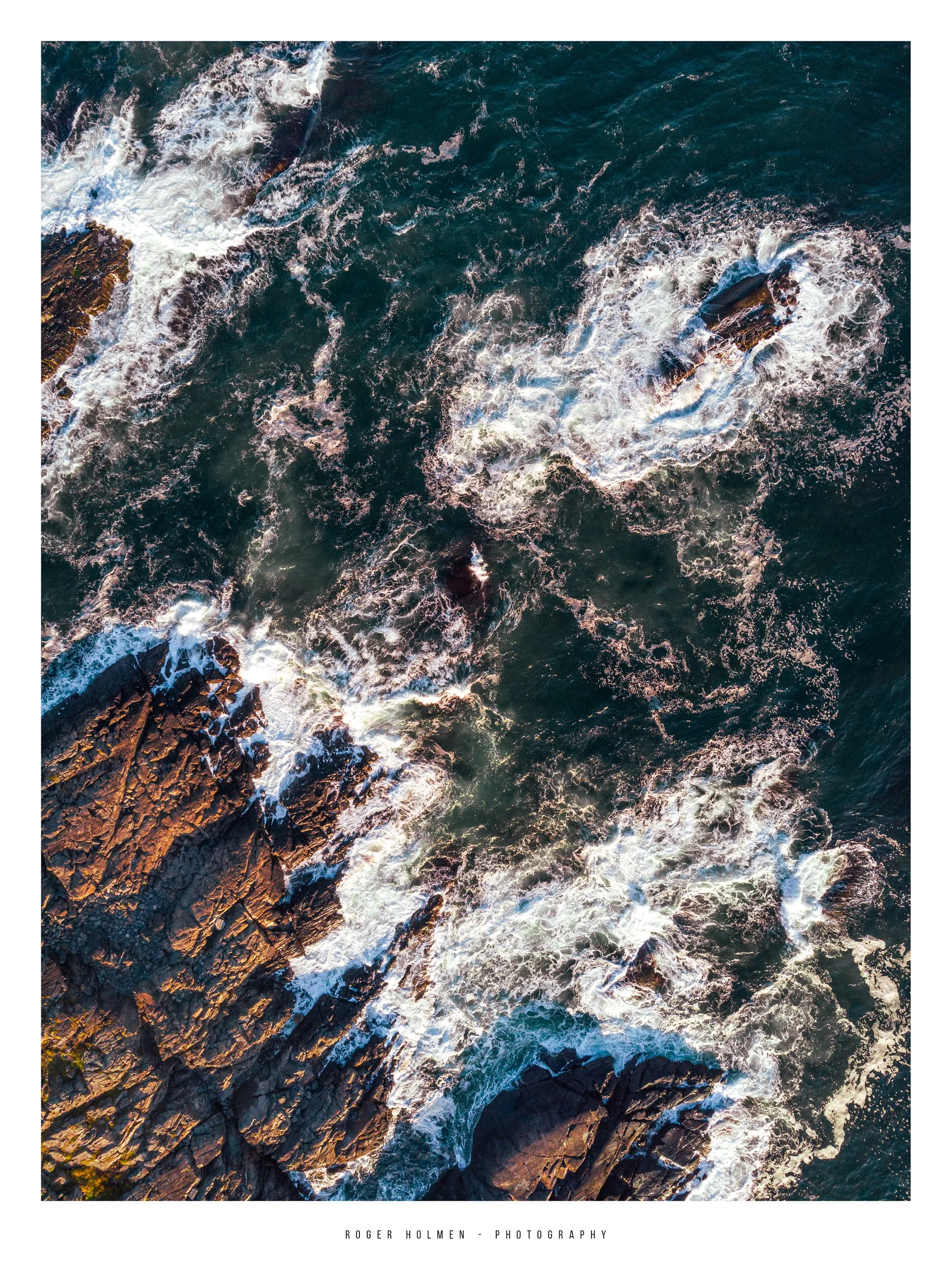 Aerial view of the ocean with waves crashing against rocky shoreline, showing white foam and a large submerged rock.