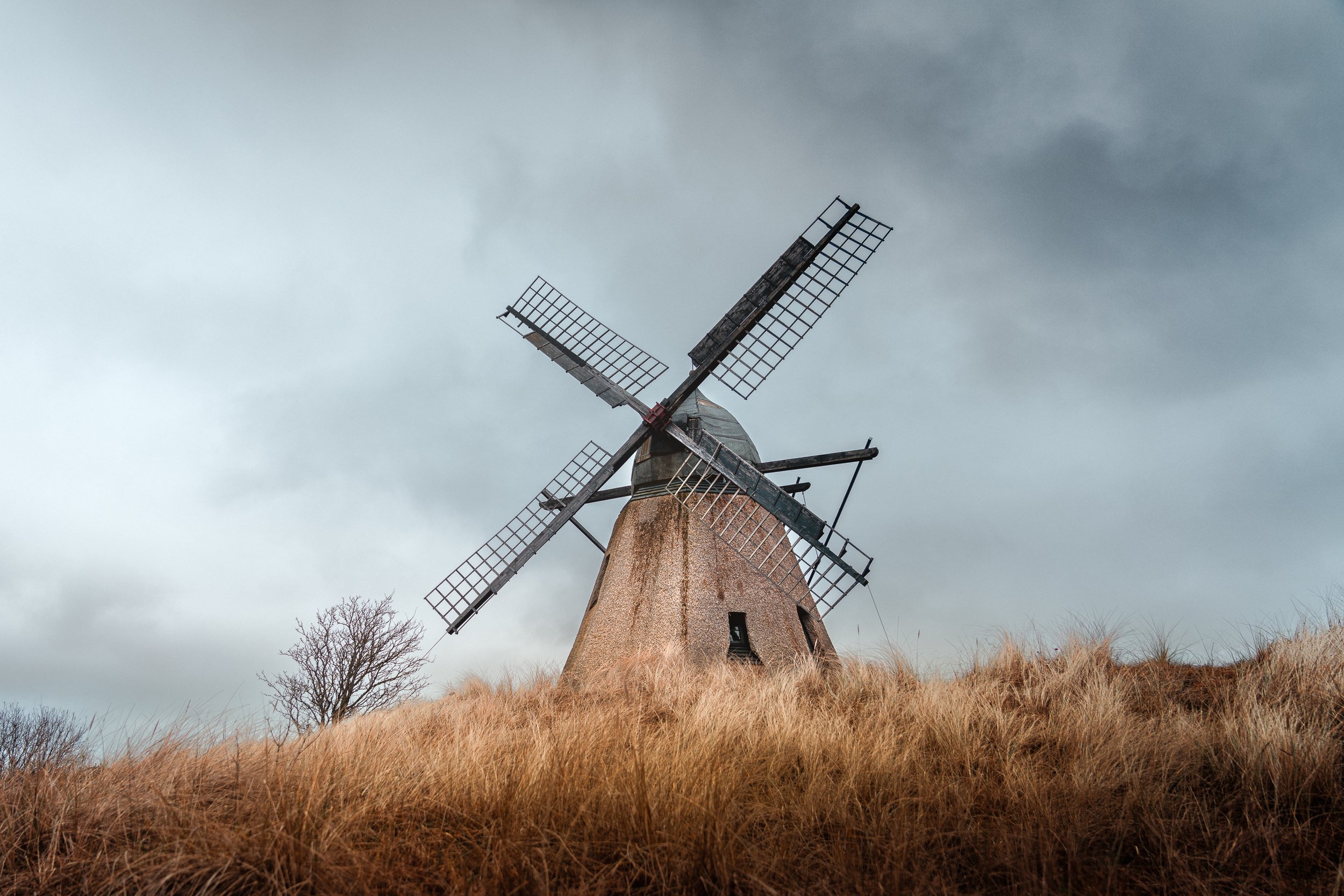 An old windmill with four deteriorated blades stands on a hill covered with dry grass under a cloudy sky.