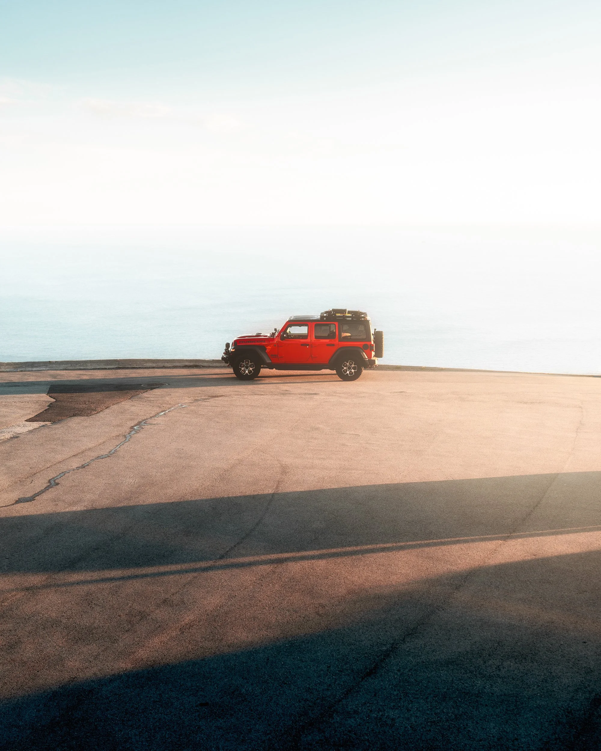 A red Jeep SUV parked on a concrete surface near a body of water with a clear sky in the background.