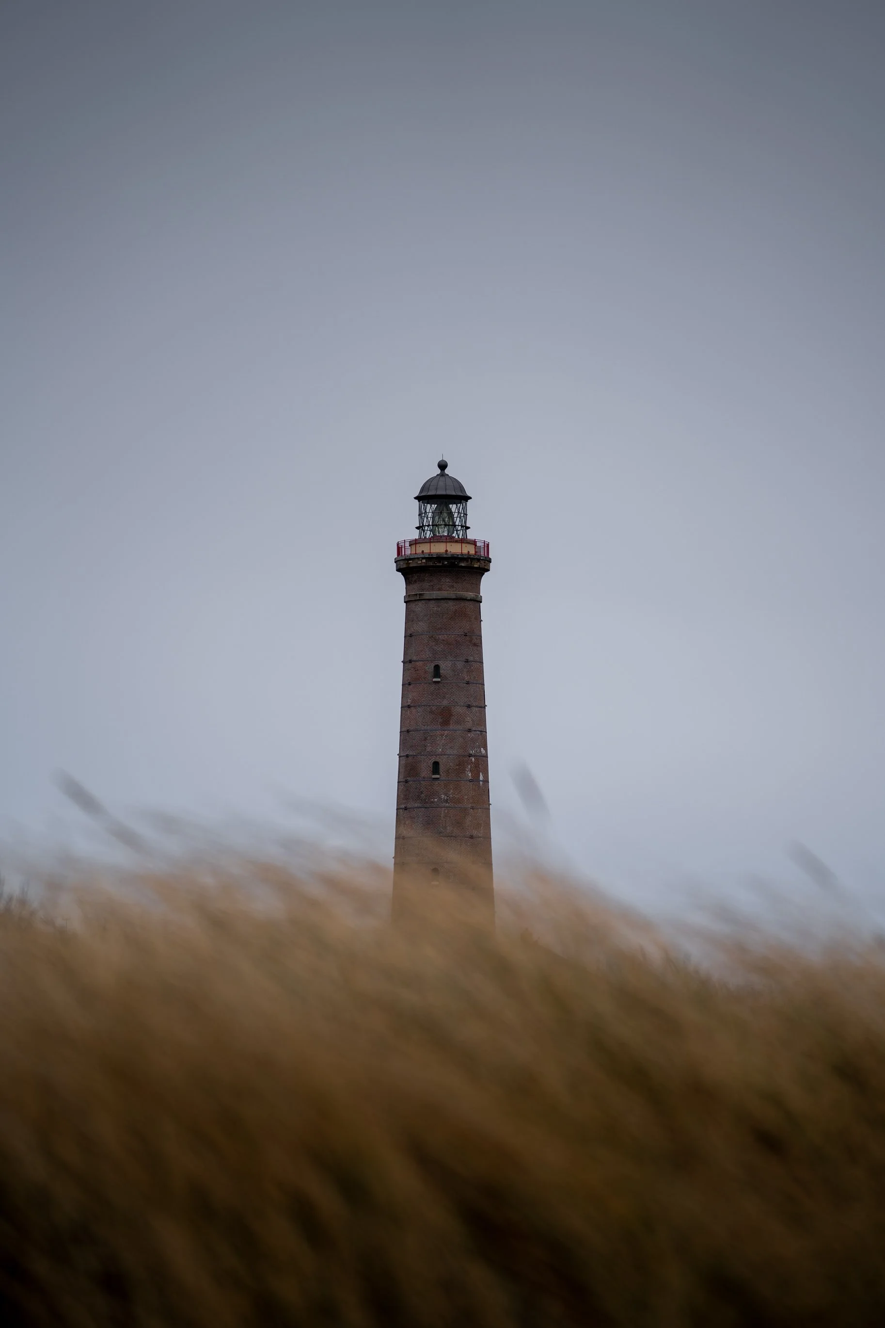 A lighthouse on a grassy hill with blurred grasses in the foreground and a cloudy sky in the background.