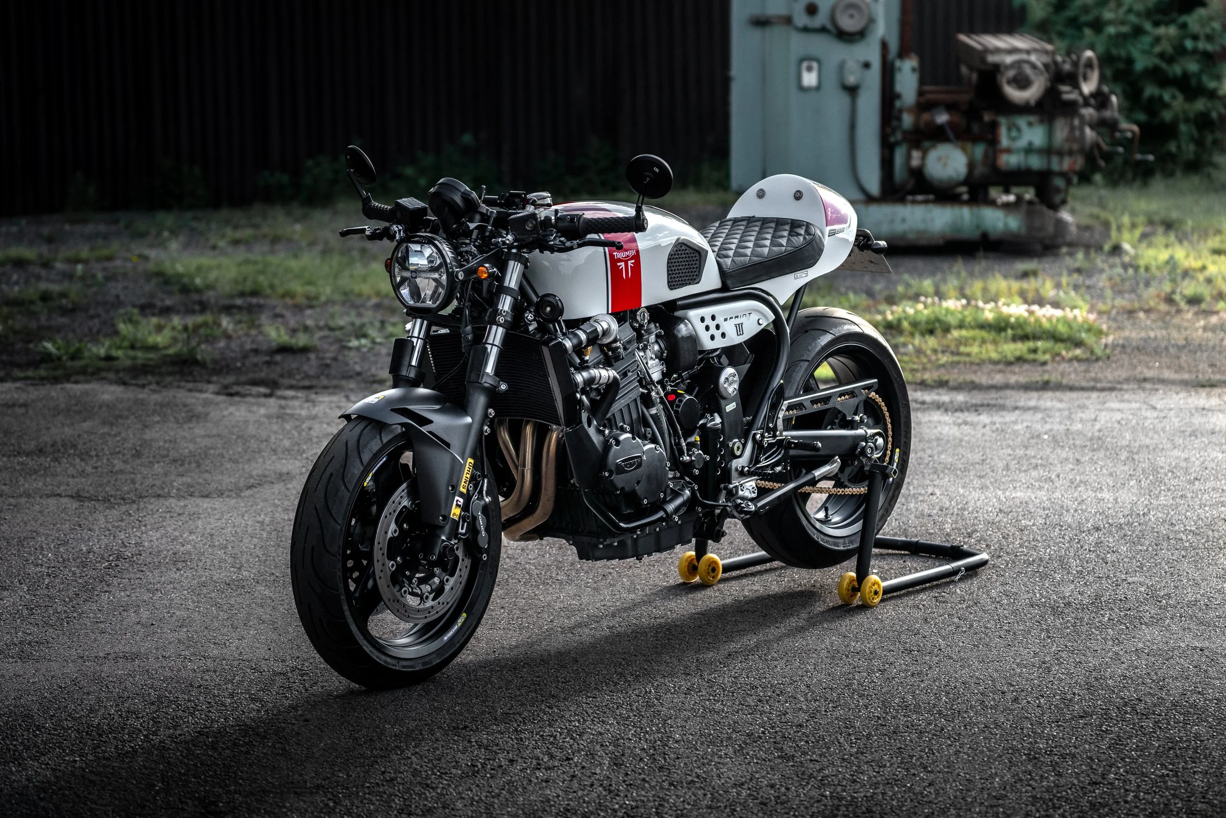 A modern custom Triumph motorcycle with a white and black color scheme, parked on a paved surface with a black fence and industrial equipment in the background.
