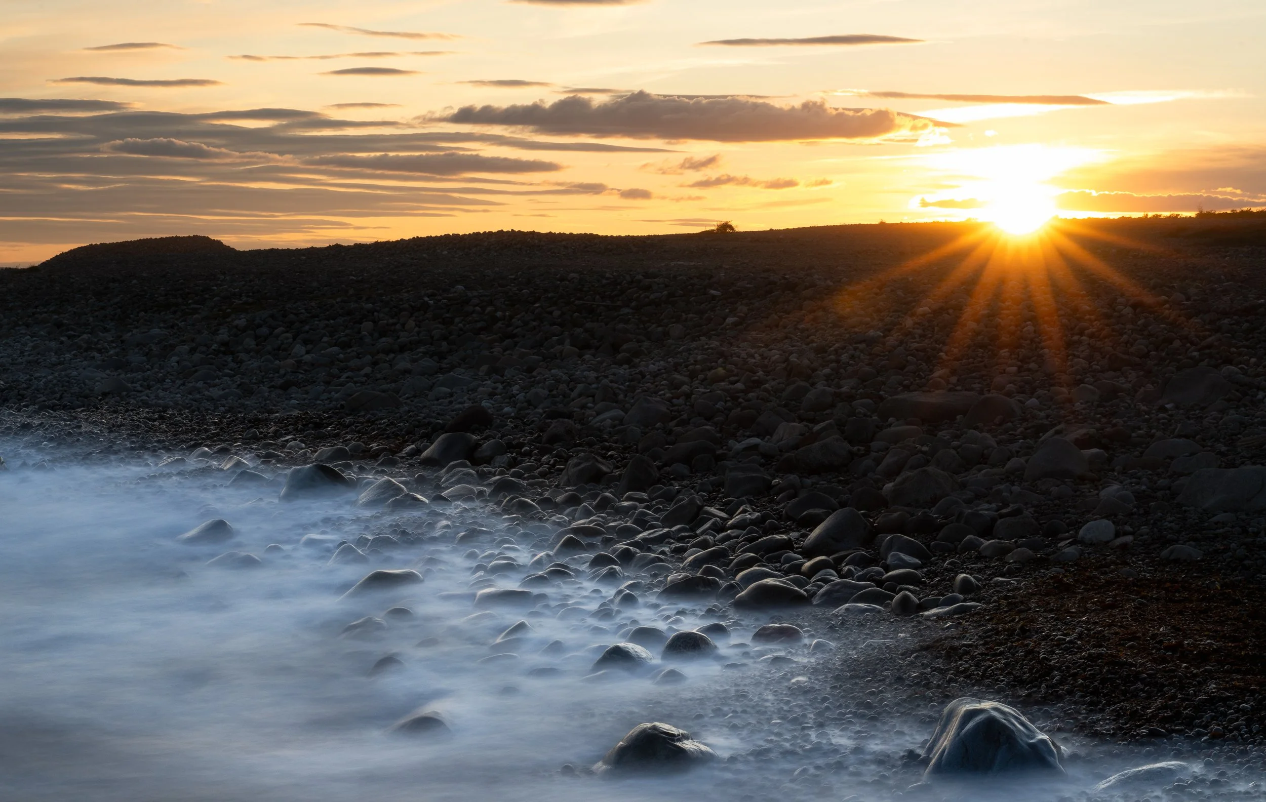 A rocky shoreline at sunset with the sun partially obscured by clouds, casting a golden light across the sky and ocean. Small waves crash against smooth stones on the beach.