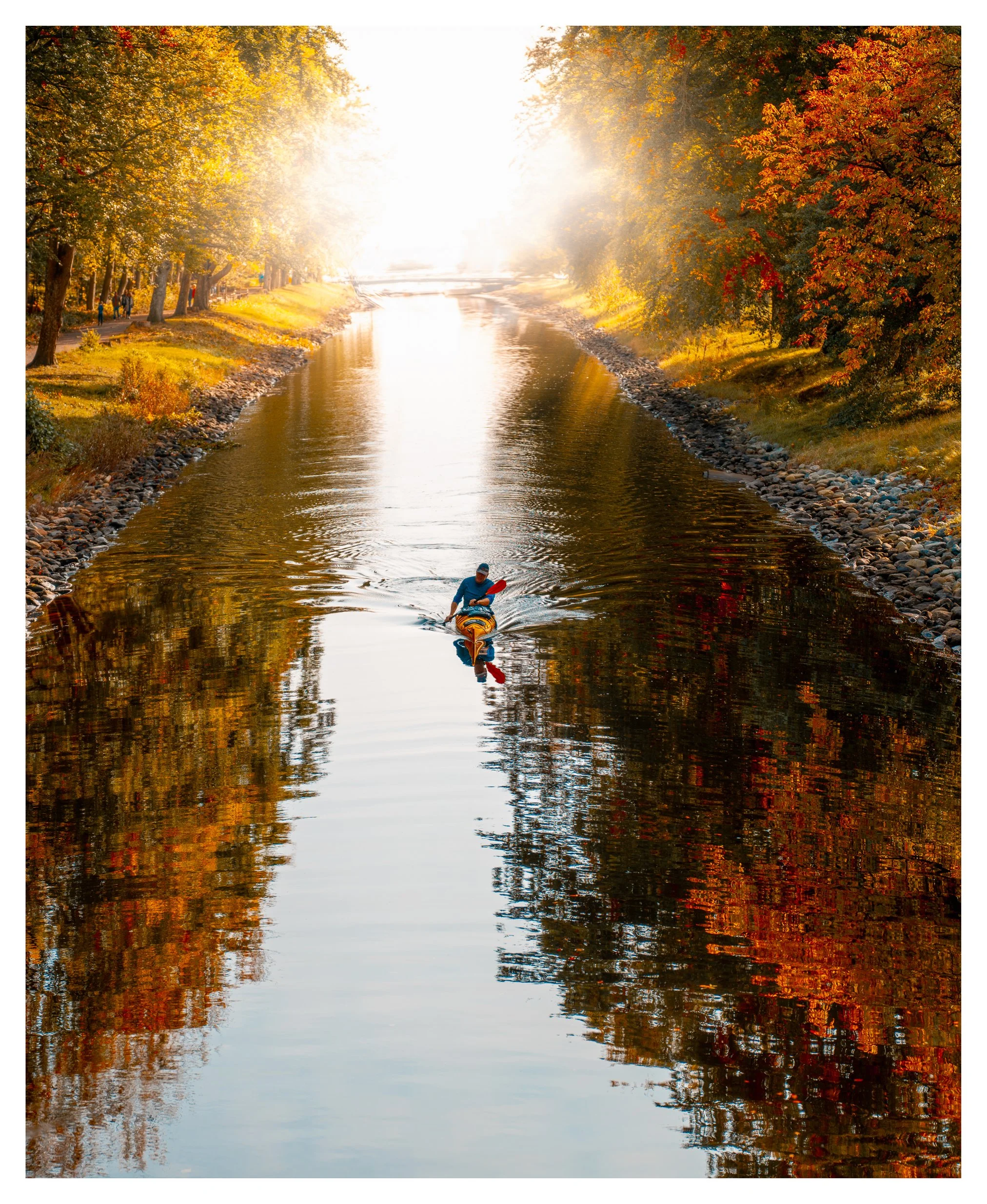 A person kayaking on a calm river during autumn, with trees displaying fall foliage on both banks and sunlight creating a misty glow in the background.