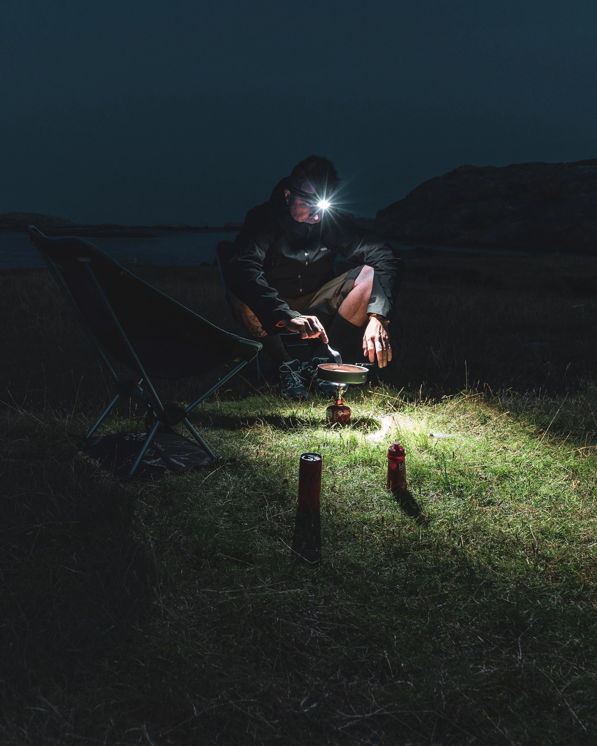 A person camping outdoors at night cooking food on a portable stove with a flashlight on their head, sitting on a chair near a grassy area with bottles and a pan.
