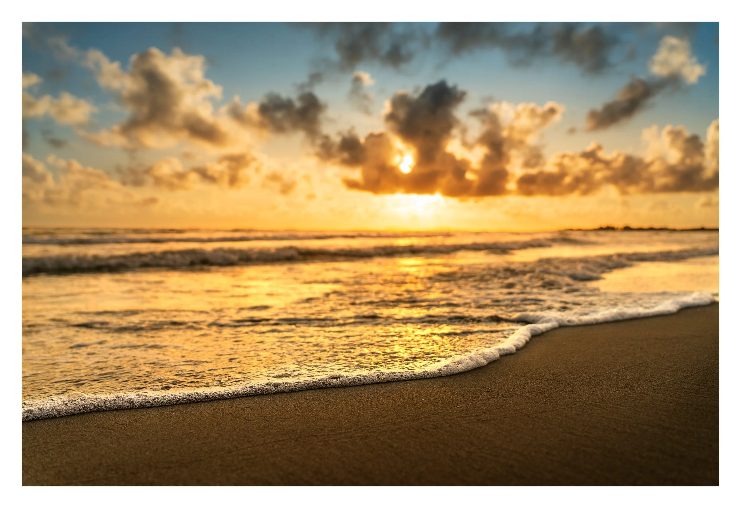 Sunset over the ocean with a cloudy sky, reflecting golden light on the water and sandy beach in the foreground.