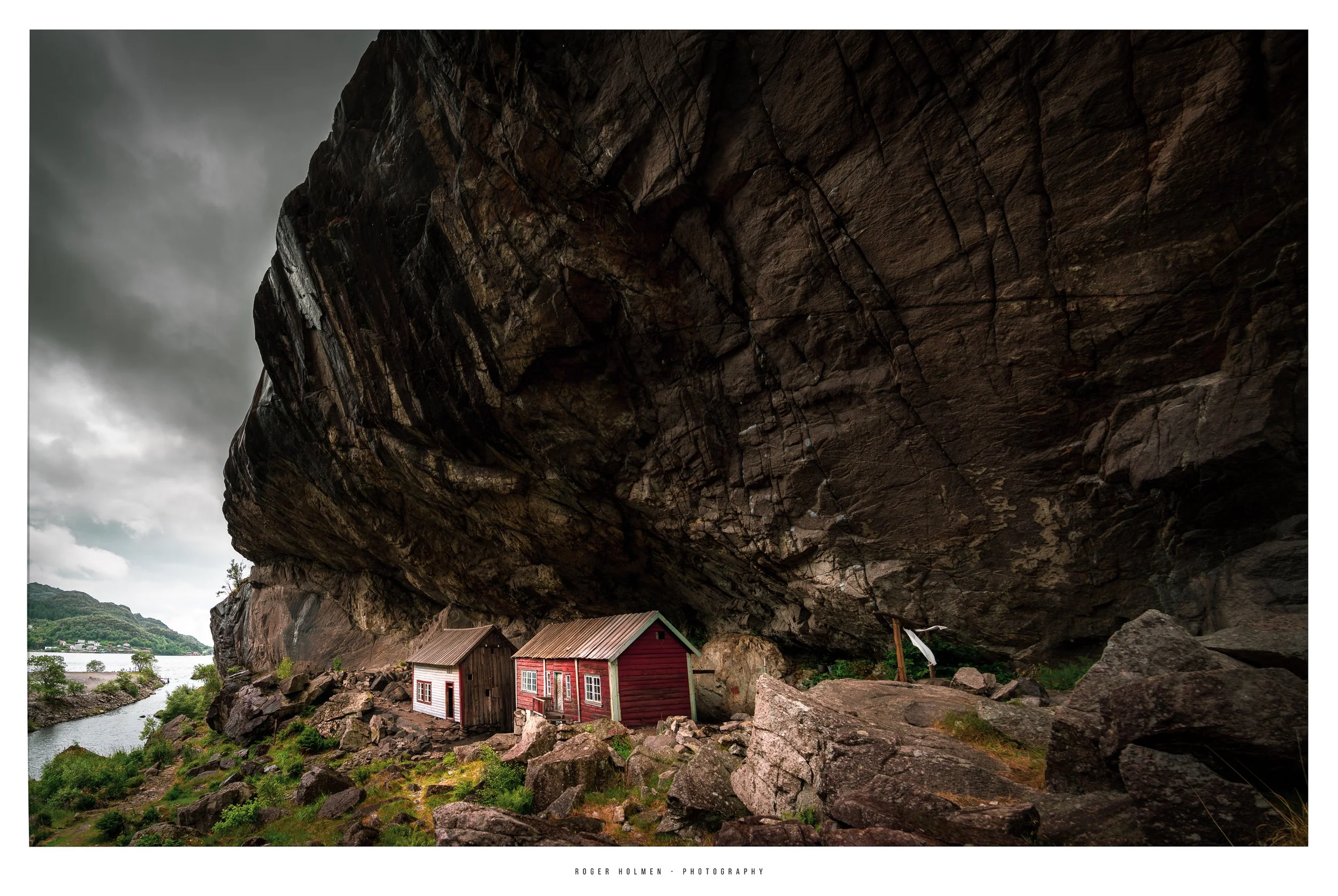 Two small houses, one red and one white, nestled at the base of a large overhanging rock formation near a river.