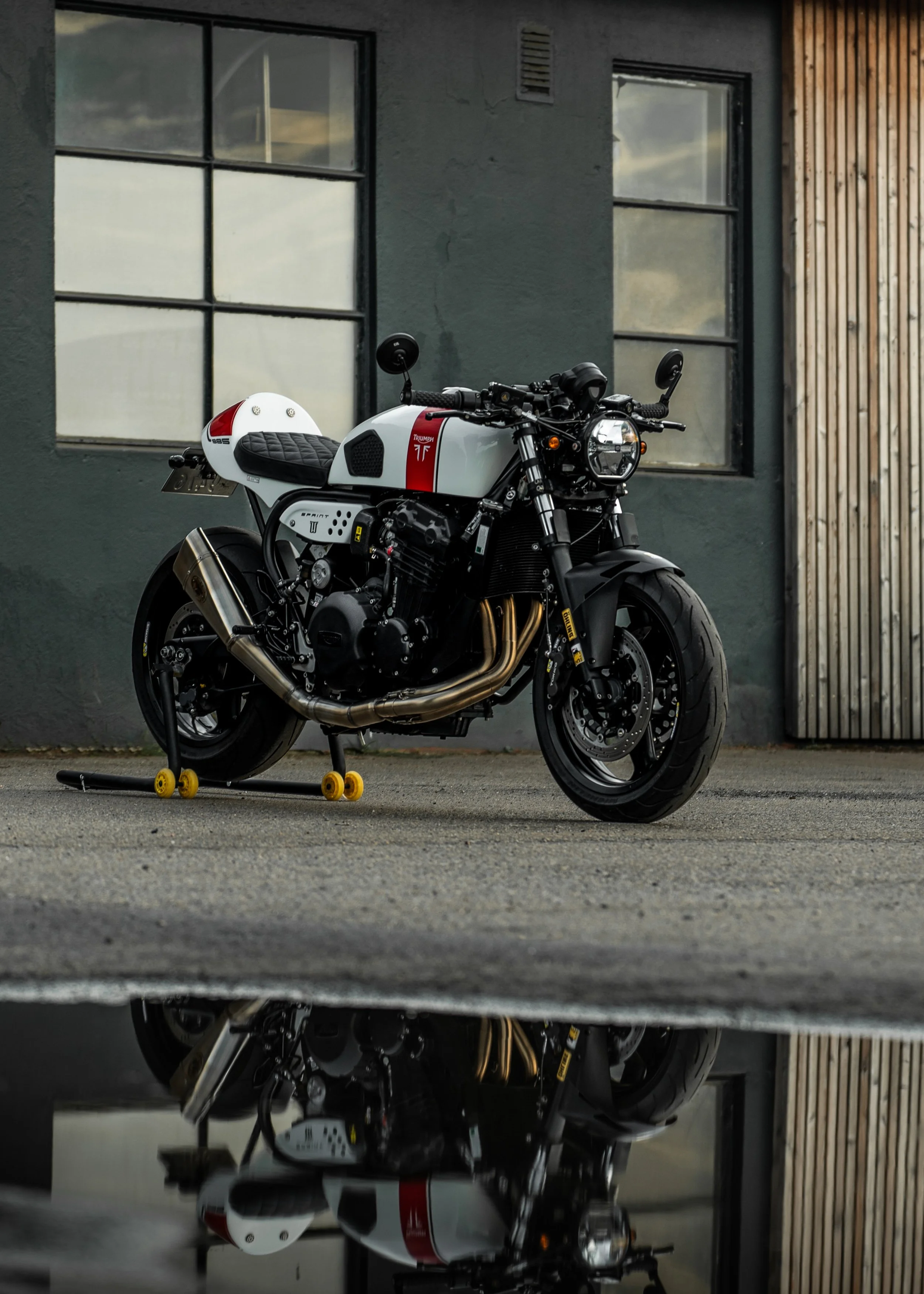 A black and white Triumph motorcycle with a red stripe and a white helmet with red accents, parked outside near a grey building with wooden panels and windows, reflected on a puddle of water.