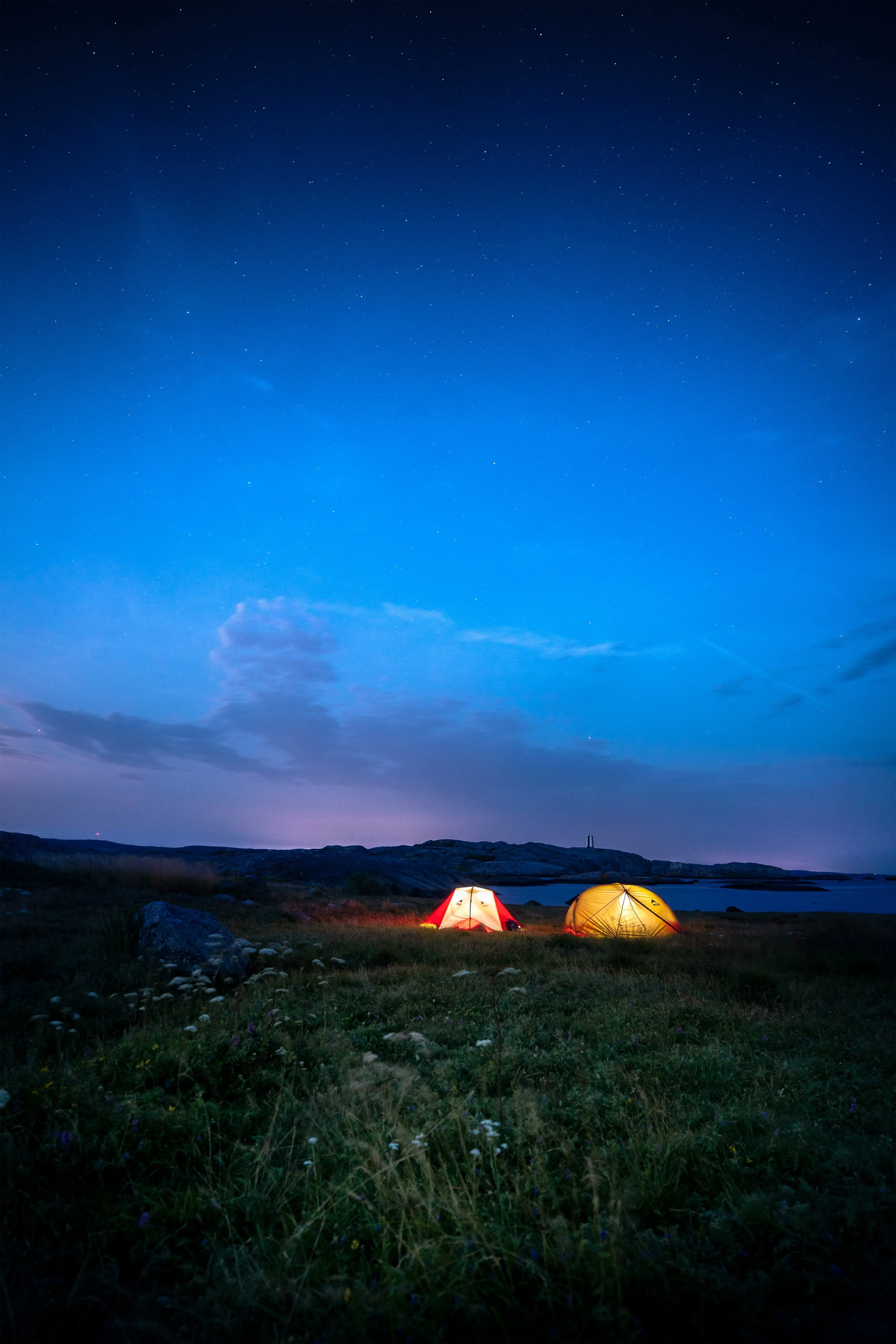 Nighttime scene of two tents illuminated on a grassy field by water, under a starry sky with some clouds.