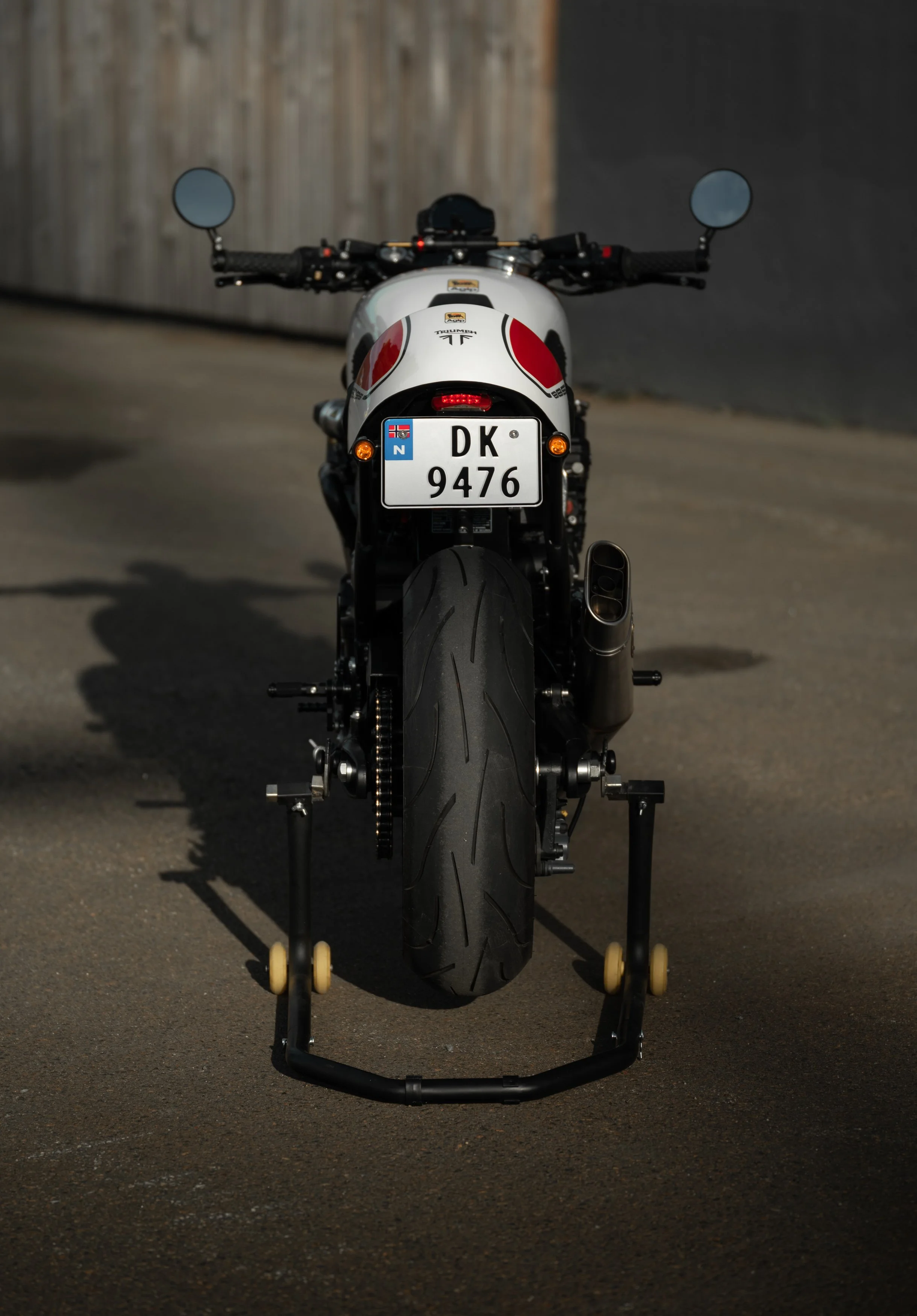 Rear view of a white motorcycle with black tire on a paved surface, showing handlebars, mirrors, and license plate, with a wooden fence in the background.
