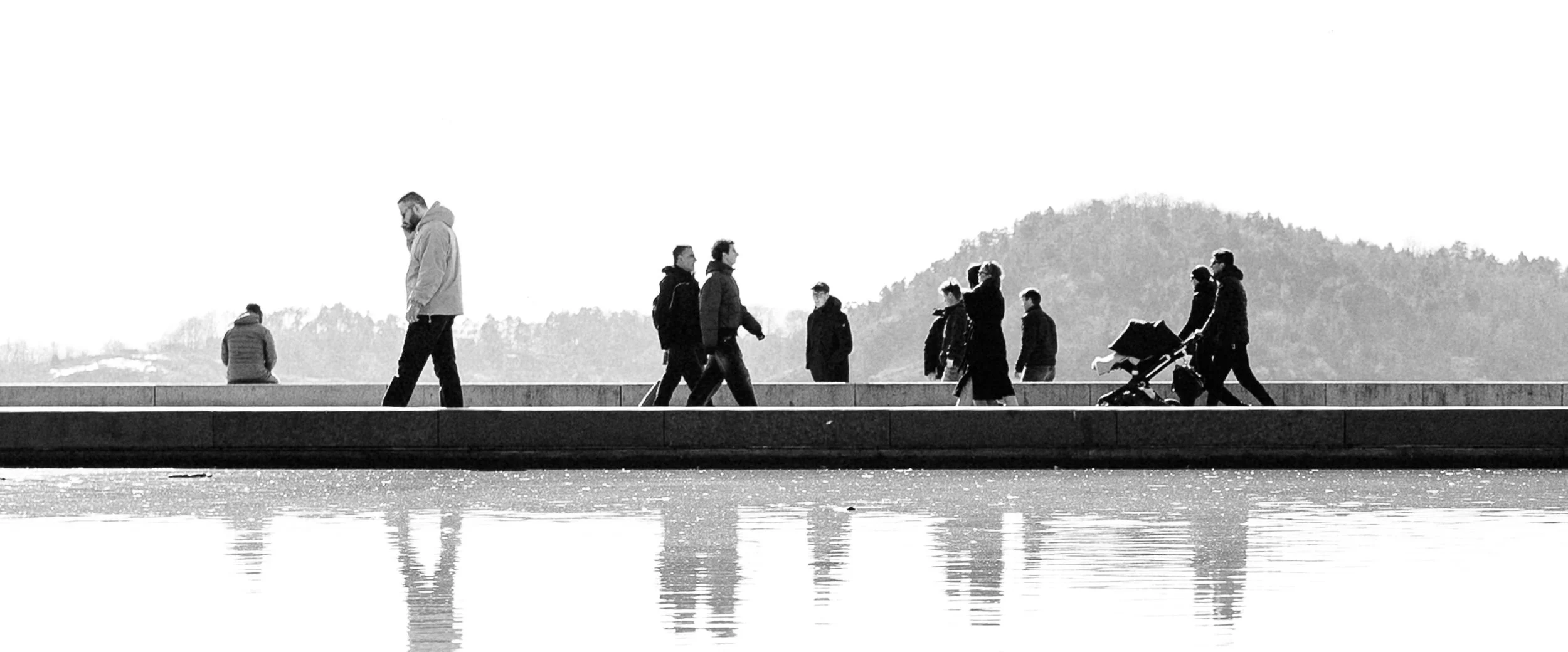 People walking along a waterfront promenade, some pushing strollers, some sitting or standing, with a mountain in the background, in black and white.