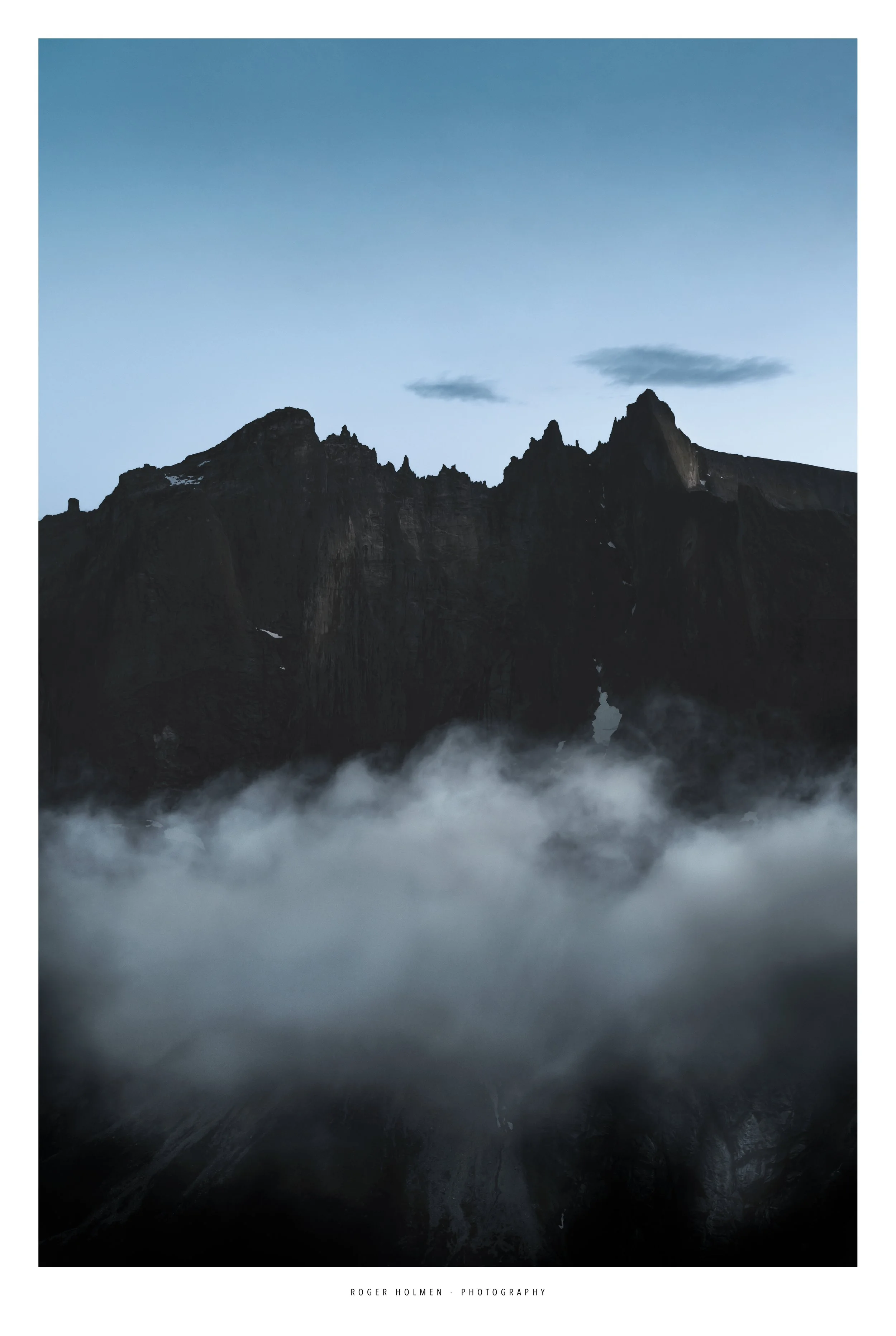 A mountainous landscape with sharp peaks, mostly dark with some patches of snow, partially obscured by a layer of fog at the base, under a pale blue sky with a few clouds. The Troll wall. Trollveggen i Romsdalen. Natur foto.