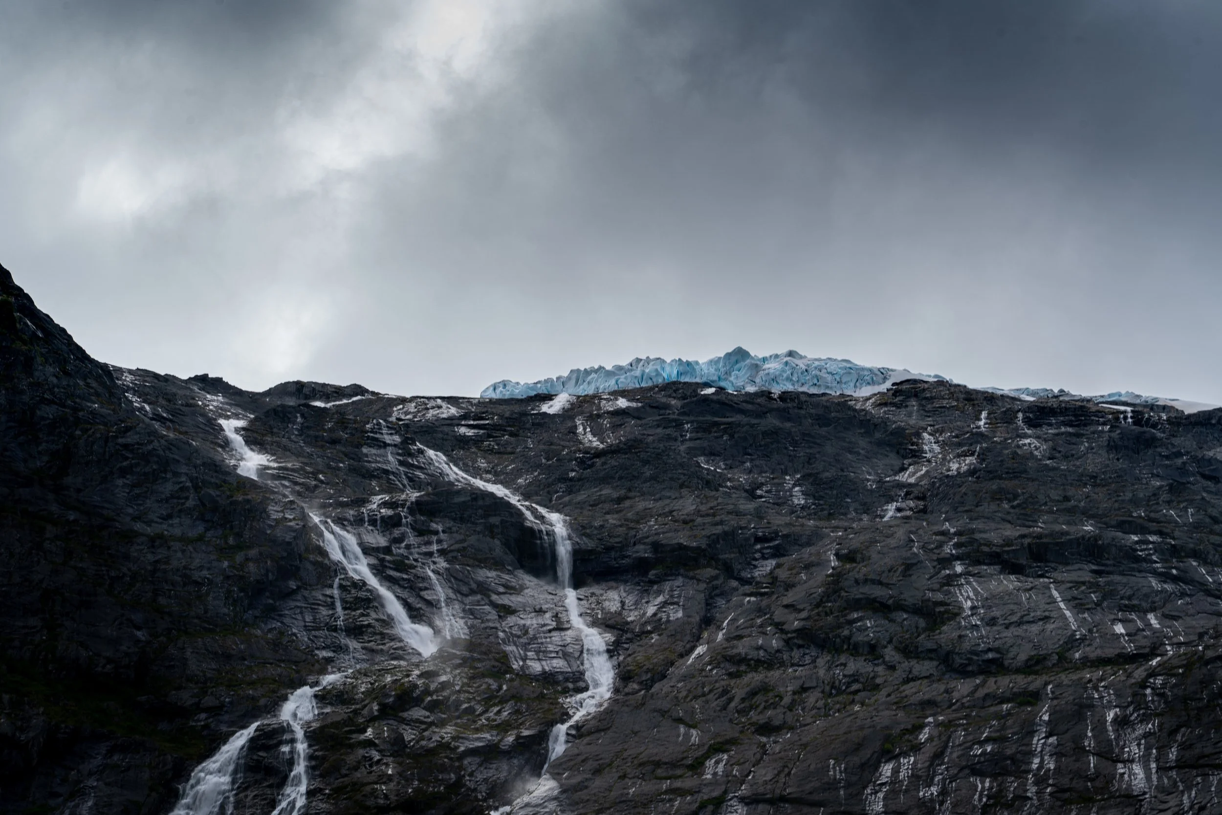 A rugged mountain with a small waterfall and a glacier in the distance under a cloudy sky.
