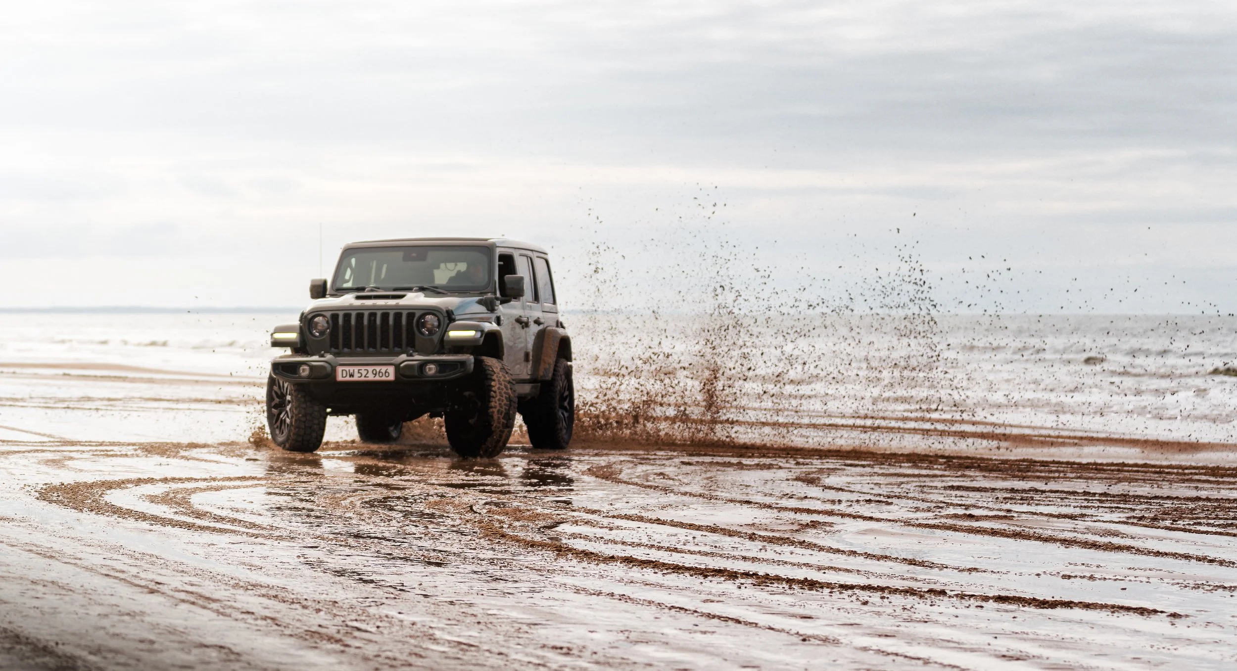 A black Jeep driving on the wet sandy beach, splashing water as it moves with cloudy sky overhead.
