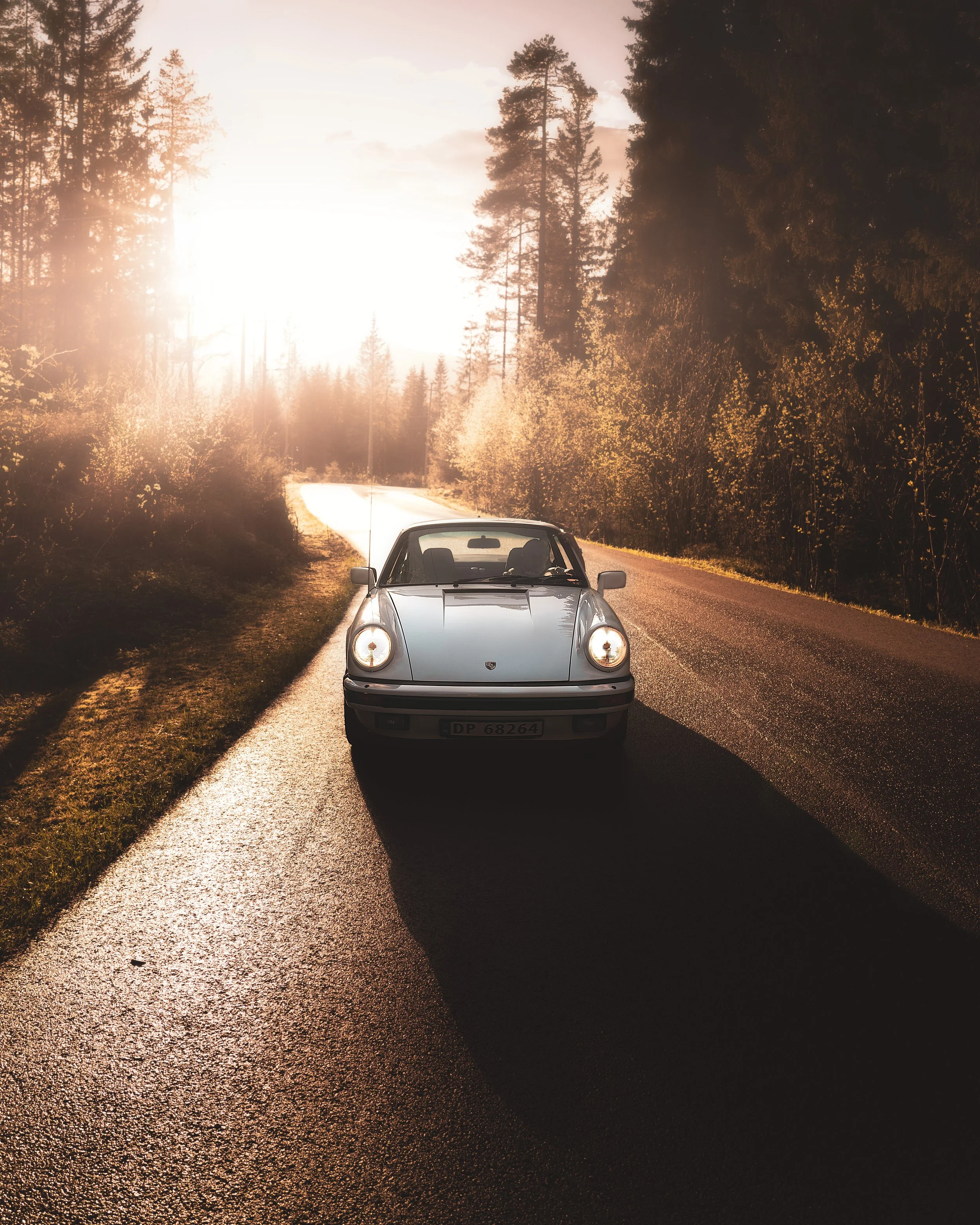 A silver sports car parked on the side of a rural road during sunset, with a forest of tall trees on either side.