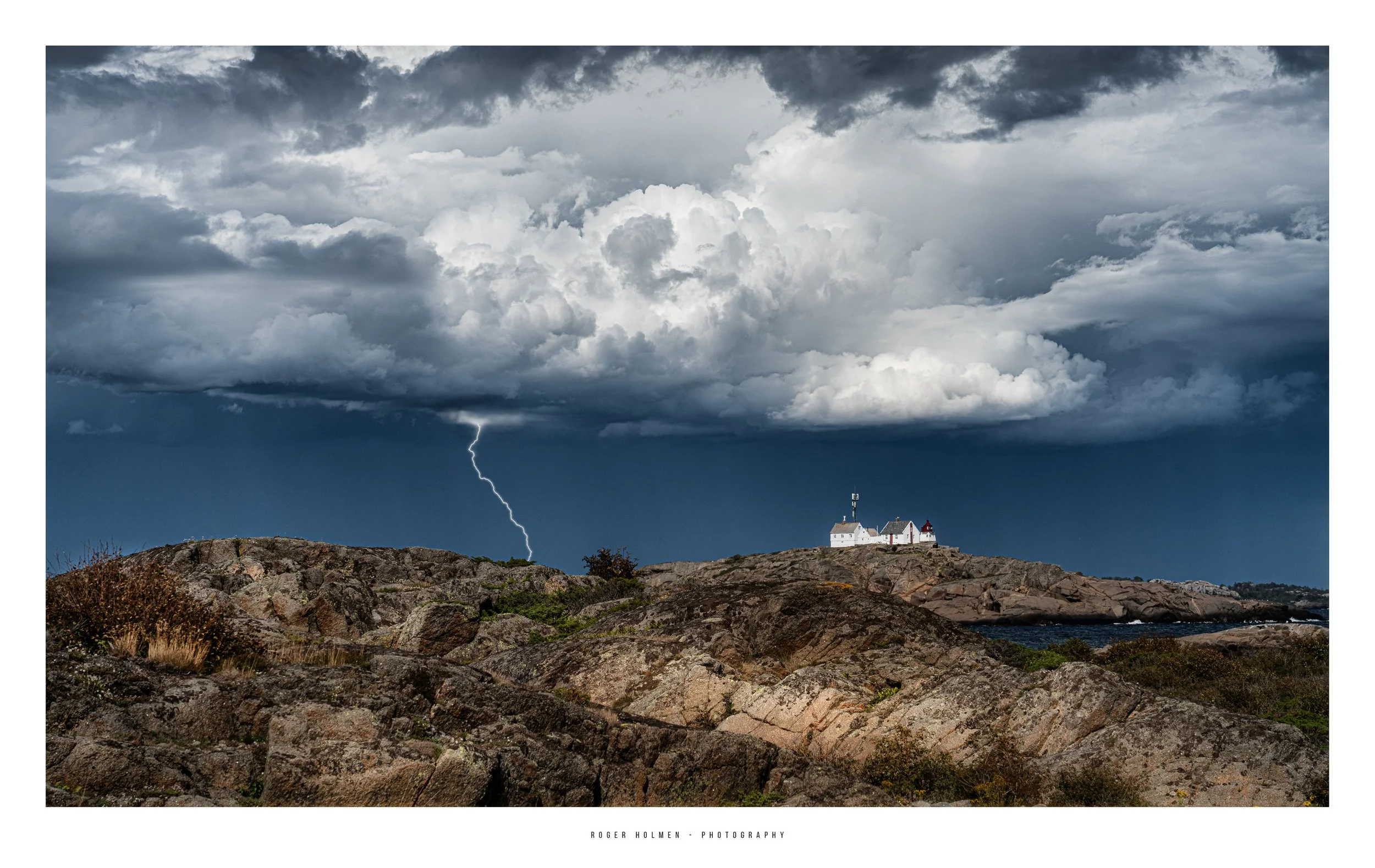 Stormy sky with dark clouds and lightning over rocky coastline and a white lighthouse with smaller buildings.