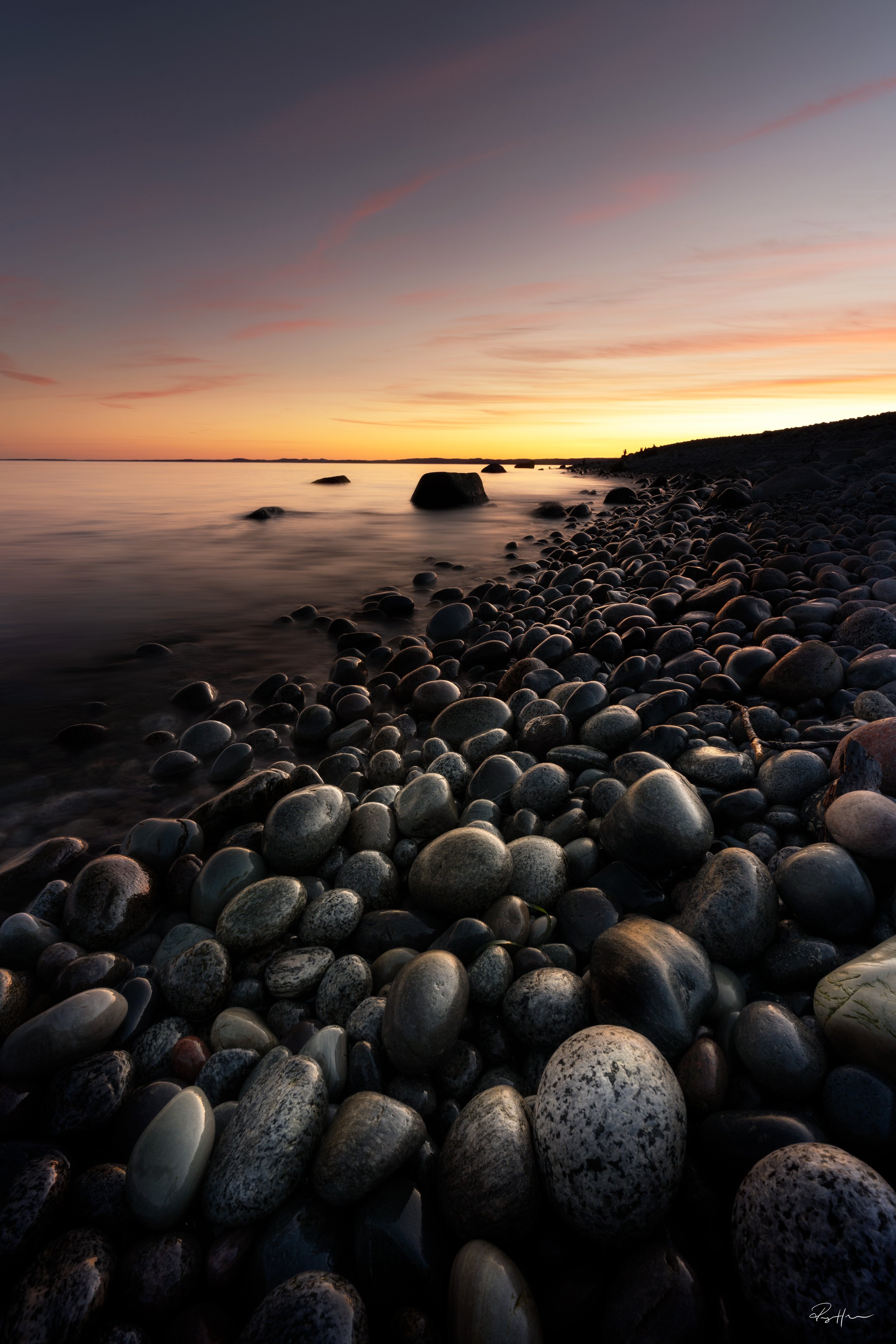 A rocky beach at sunset with smooth, dark stones in the foreground, calm water, and a colorful sky at dusk.