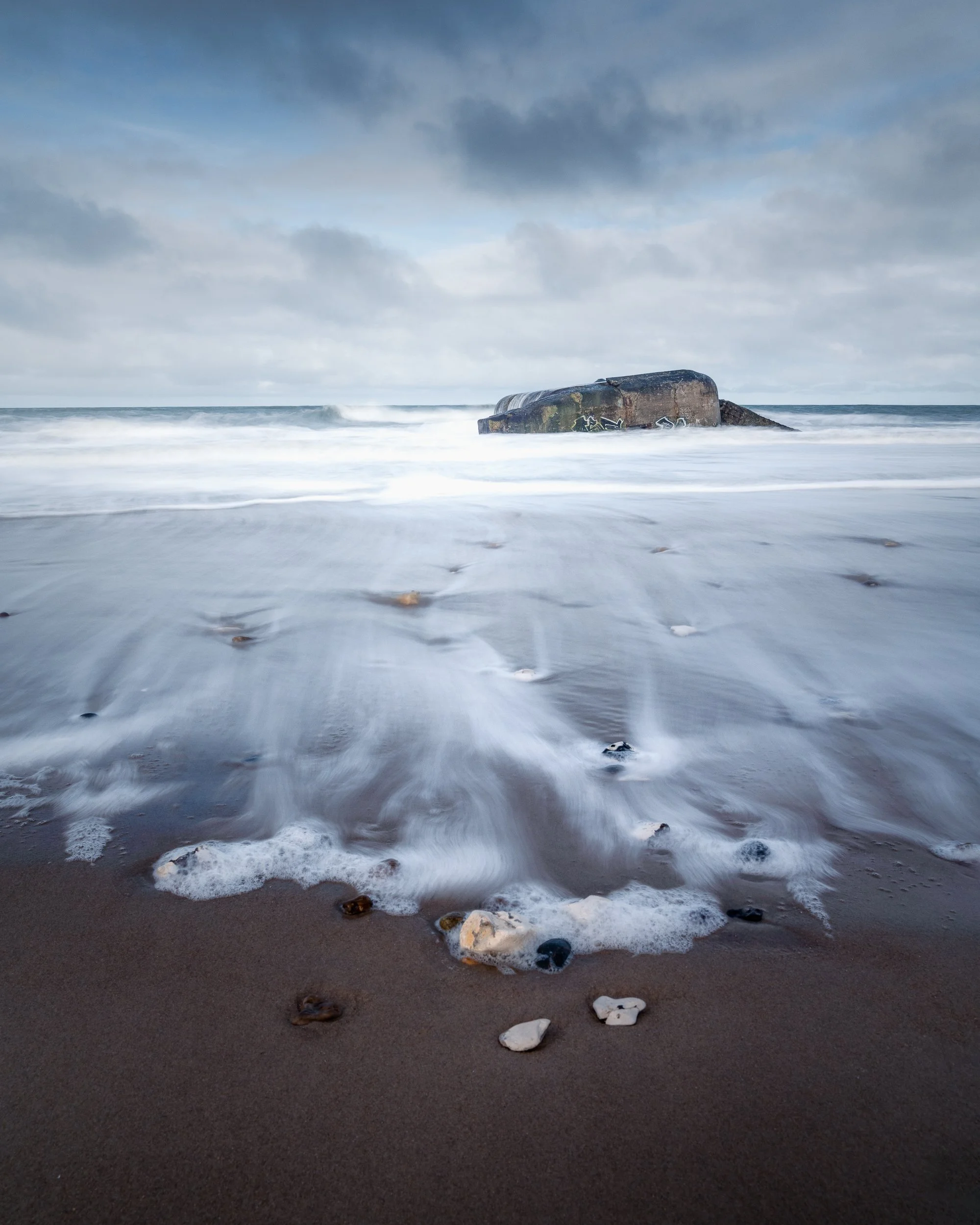 Overcast sky at a rocky beach with a large submerged object and waves washing over the sand.