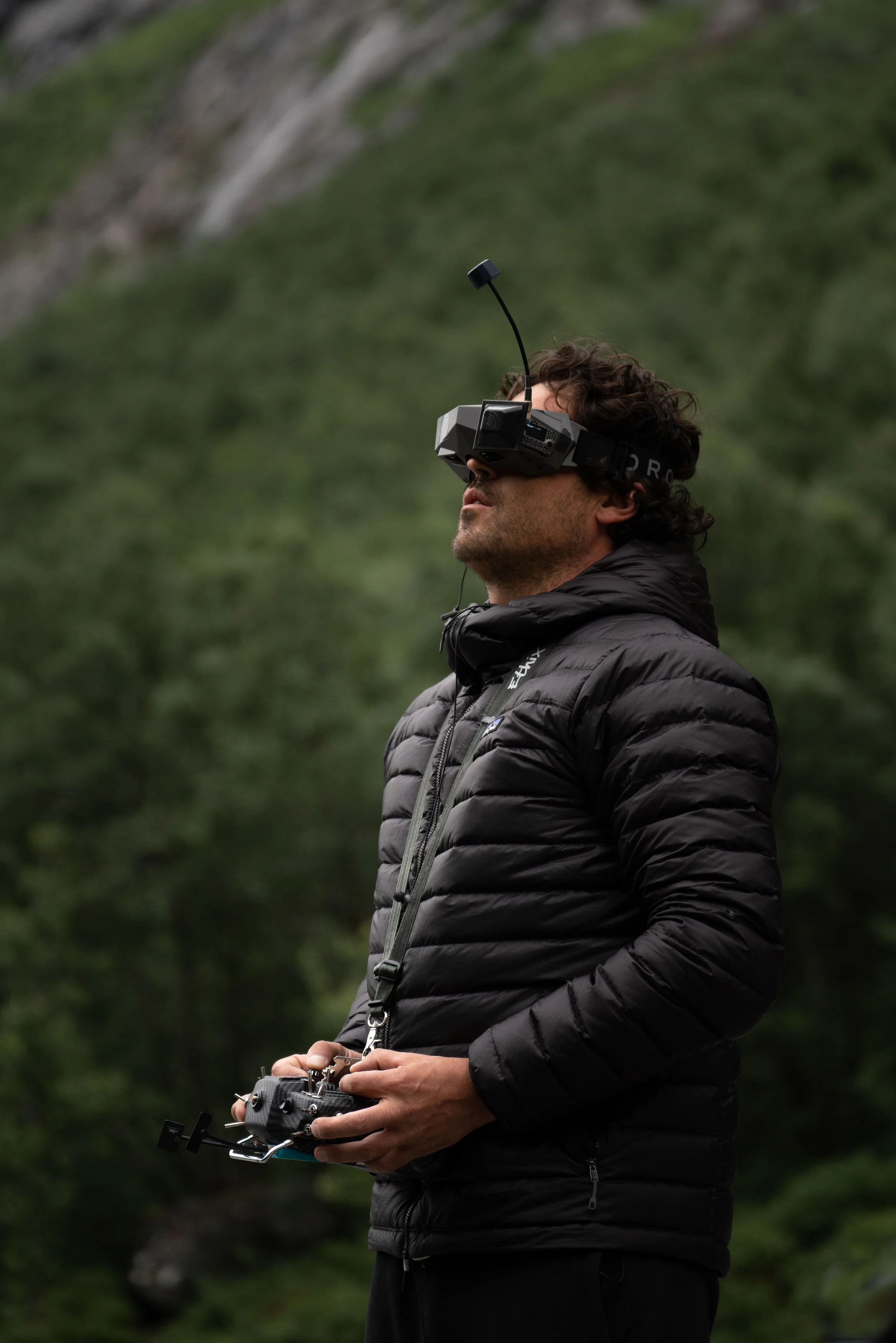 A man wearing a black jacket and goggles is outdoors, holding a remote control and flying a drone in a green, mountainous area.