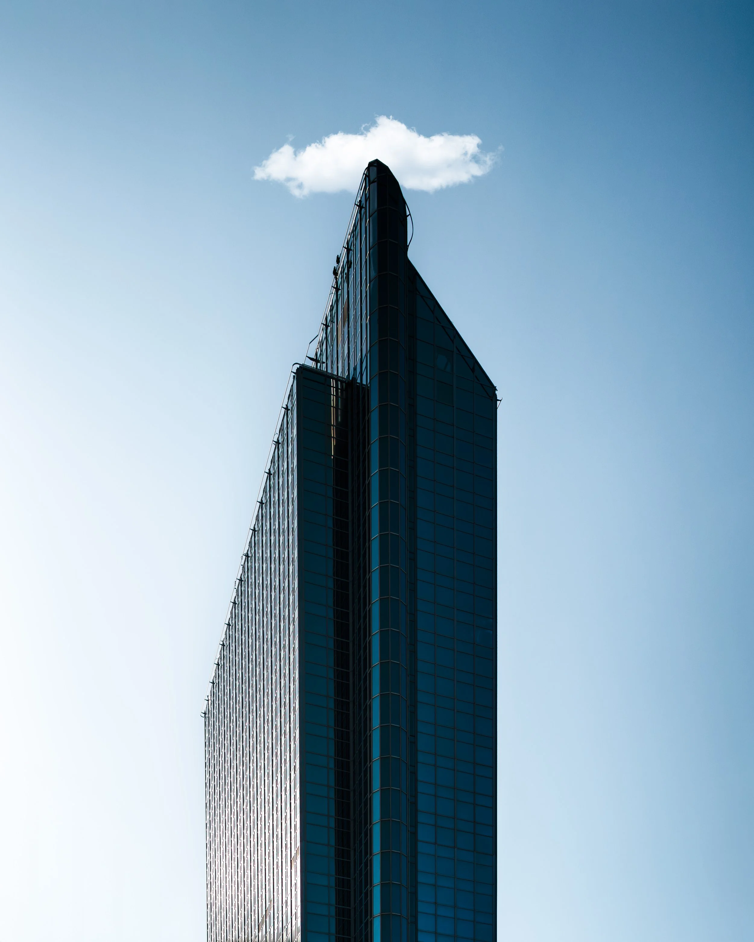 A tall modern glass skyscraper with a single cloud above it against a clear blue sky.