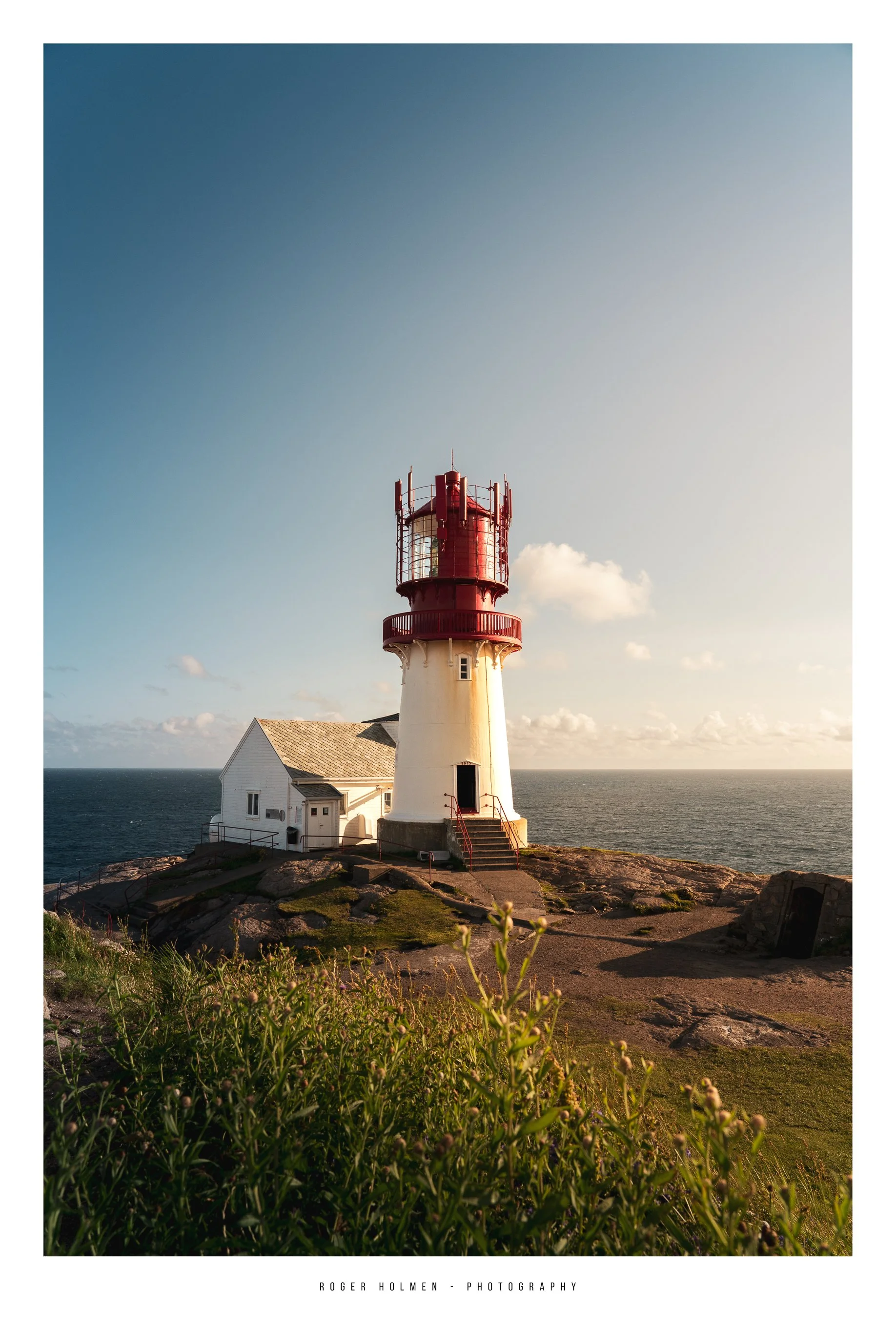 A white lighthouse with a red top and railing, adjacent to a small white building, situated on a rocky coastline with green vegetation, under a clear blue sky with clouds.