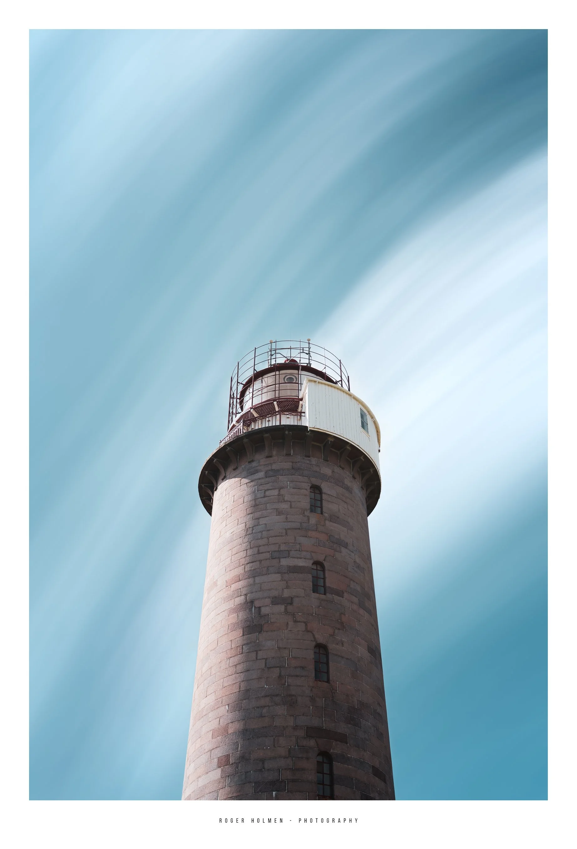 A lighthouse with a tall stone tower and a white lantern room at the top, set against a blue sky with wispy clouds.