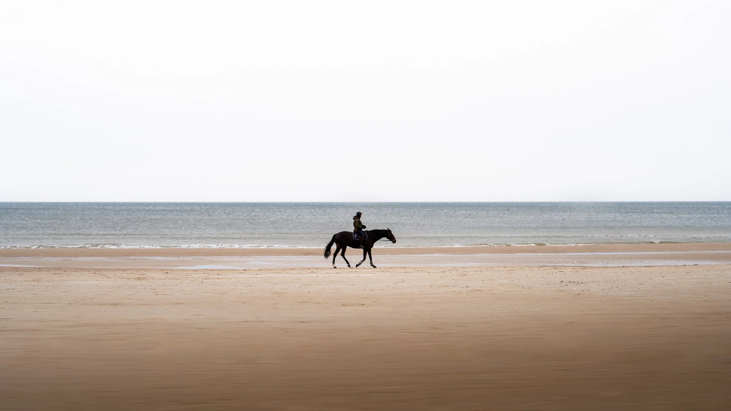 A person riding a horse along a beach with the ocean in the background and an overcast sky.
