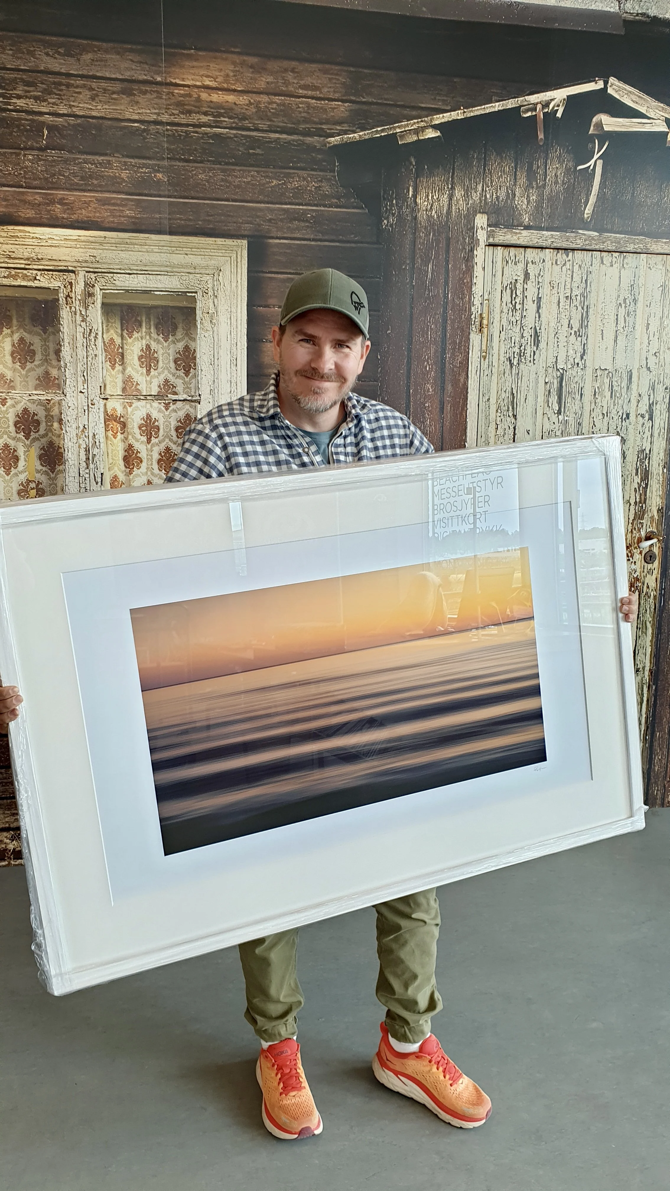 A man wearing a green cap, plaid shirt, and beige pants is holding a large framed photograph of a sunset over a body of water, with orange and purple hues.