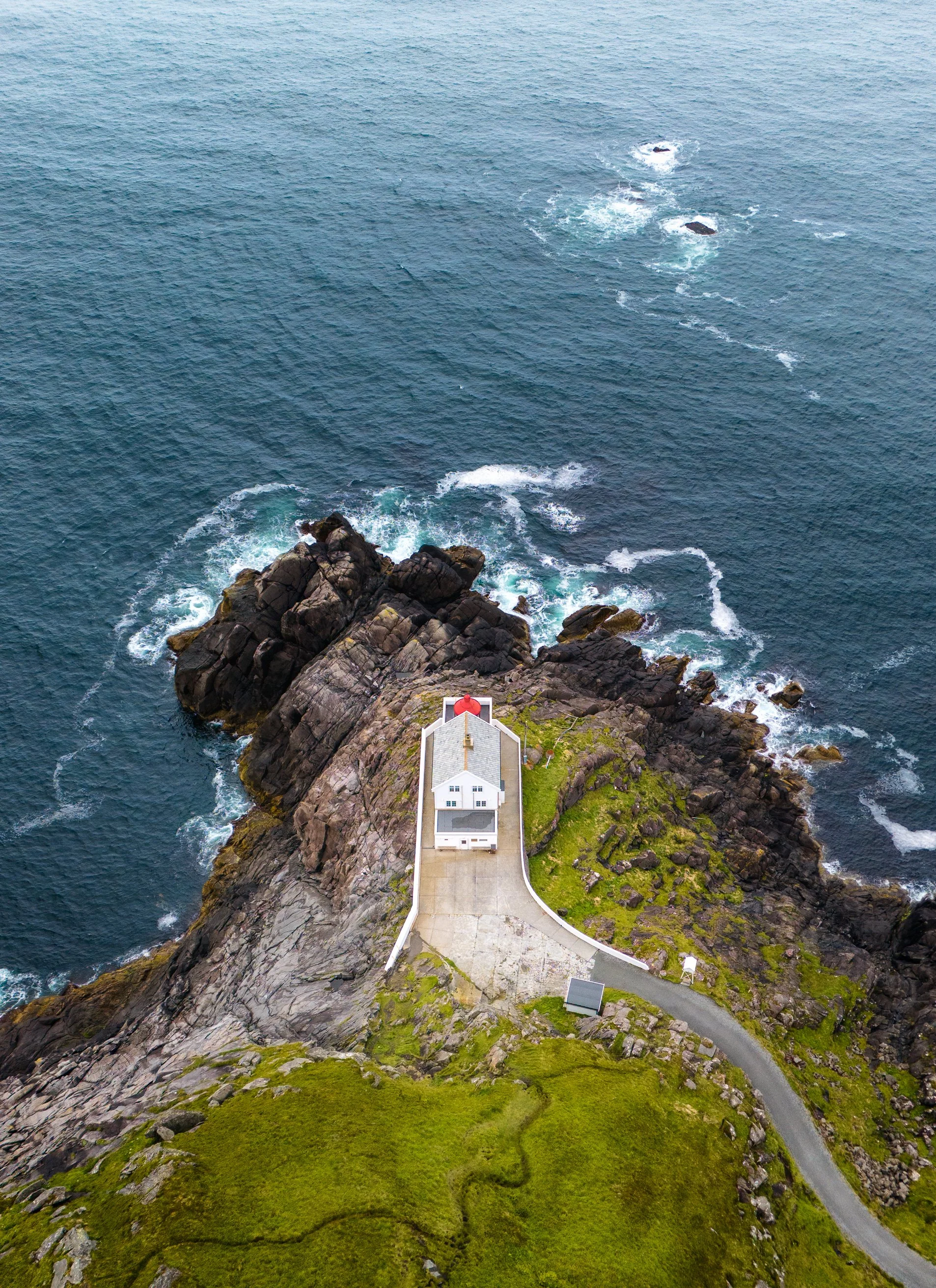 A lighthouse on a rocky outcrop at the edge of the ocean, with a winding road leading to it, surrounded by grassy land.