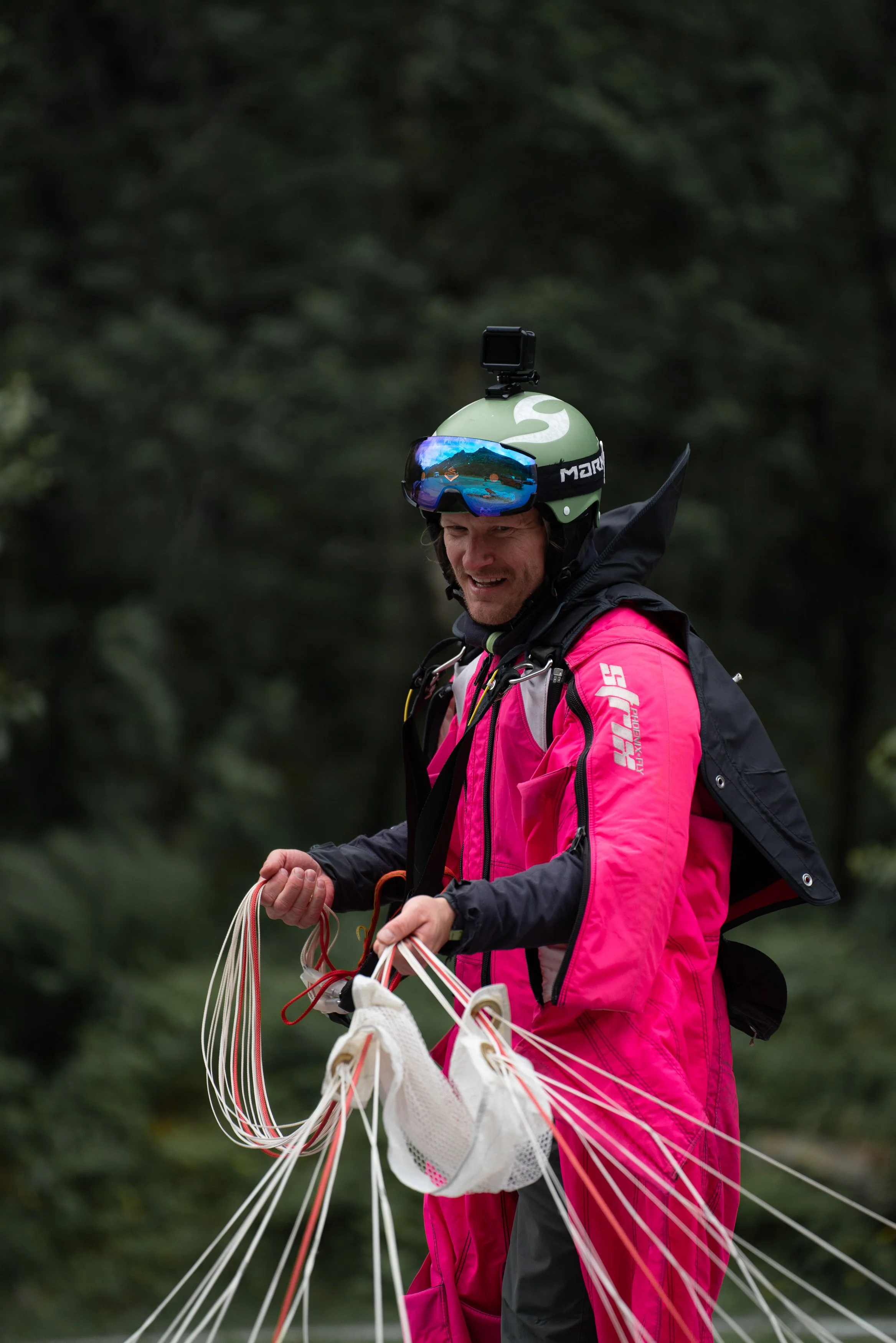 Wingsuit flyer preparing equipment outdoors in a forested area, with a helmet, goggles, and a pink jumpsuit.