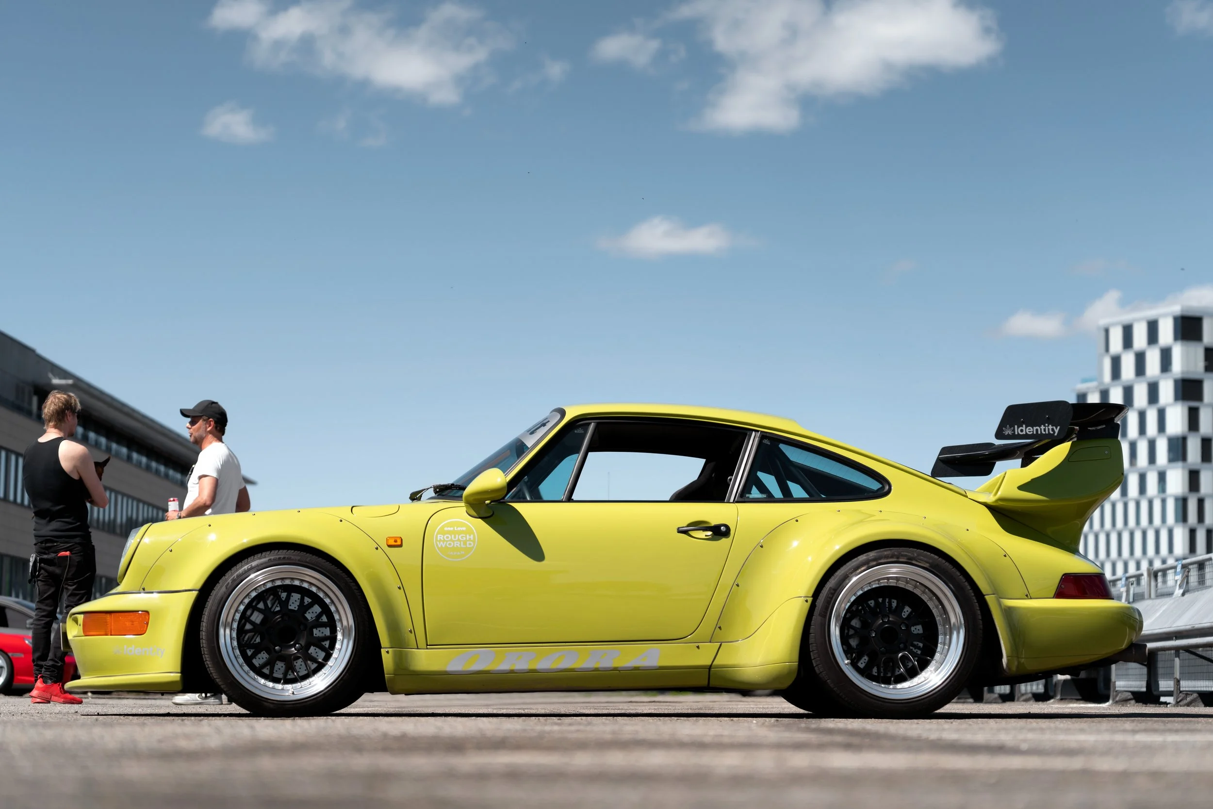 A bright yellow race car parked outdoors in a city setting on a sunny day with buildings in the background. Two men are standing nearby, engaged in conversation.