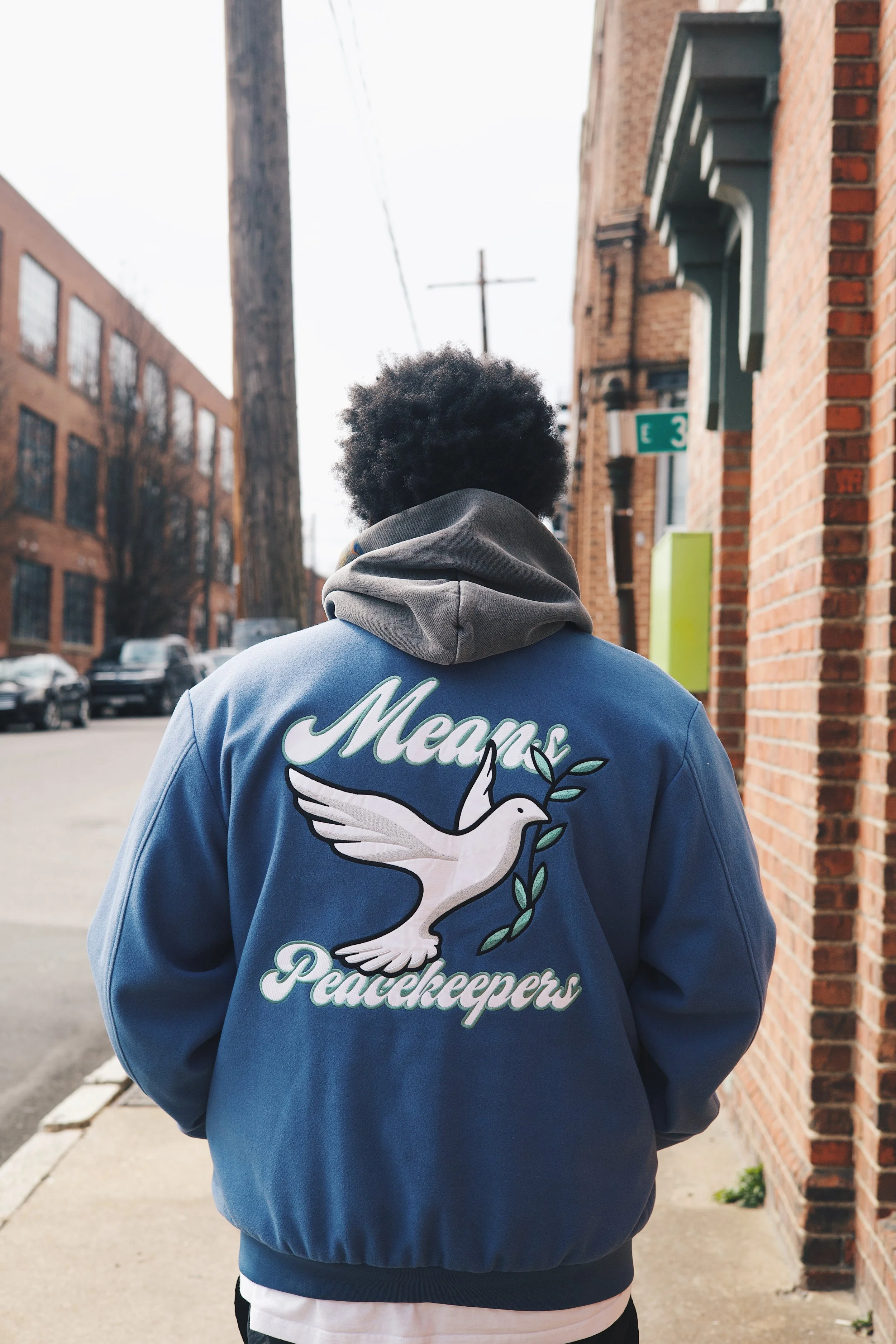 Man standing on sidewalk outdoors wearing a blue jacket with a white dove and the words 'Means Peacekeepers' on the back, with a brick building and parked cars in the background.