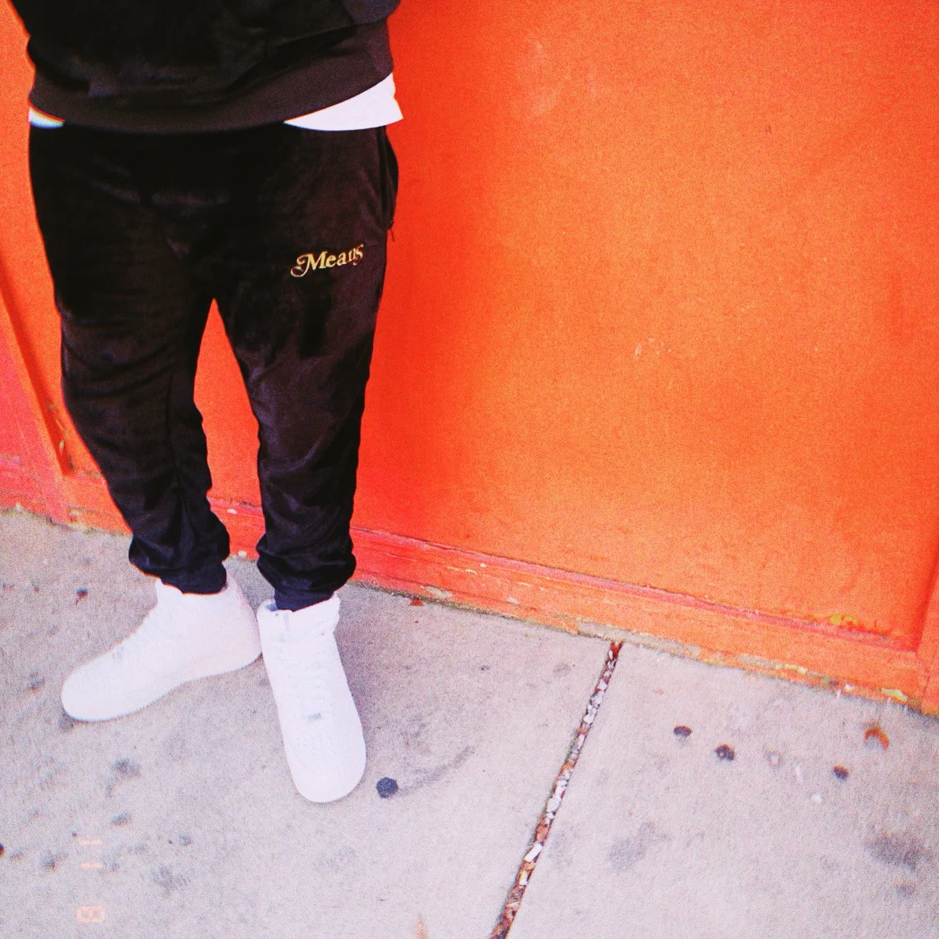Person wearing black sweatpants with gold embroidery and white sneakers, standing next to an orange wall.