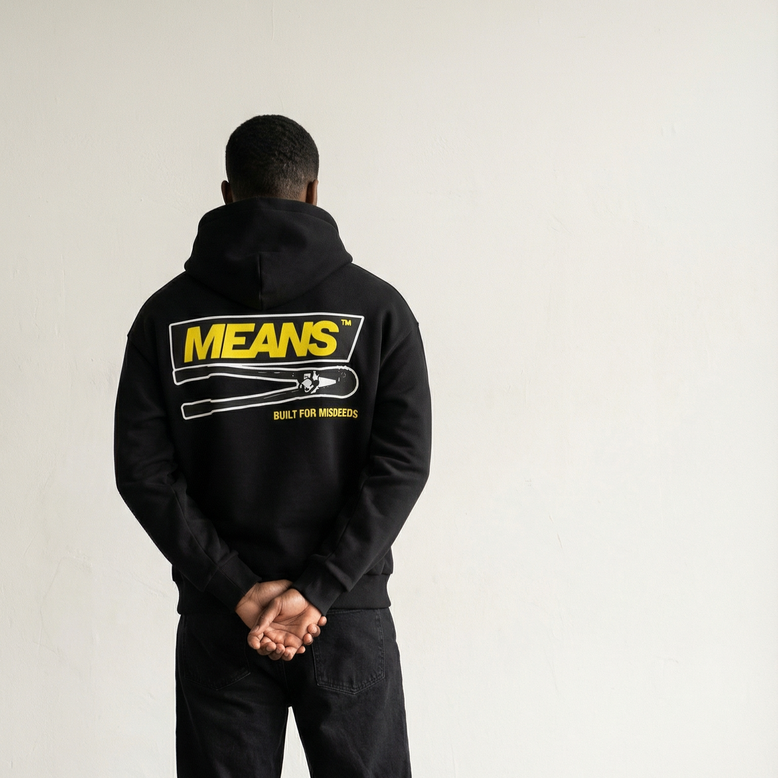 Person standing with back facing camera, wearing black hoodie with bold yellow text "MEANS" and a graphic of a hand holding a wrench, and the words "BUILT FOR MISDEEDS" underneath, against a plain white wall.