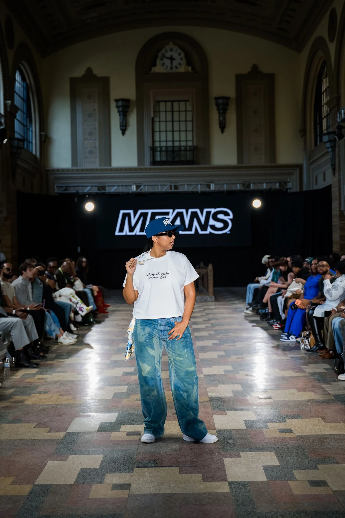 A fashion runway show in a historic building with high arched ceilings, ornate architectural details, and a large digital display with the word 'MEANS.' A model wearing baggy, tie-dye jeans, a white T-shirt with text, a blue cap, and sunglasses walks on the runway, surrounded by seated audience members.